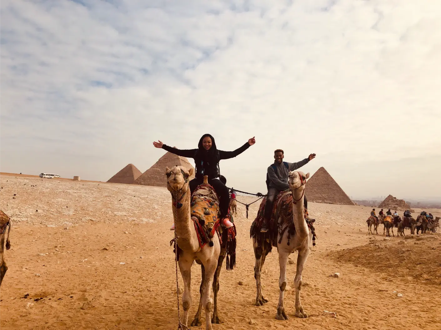 Two people riding camels with the pyramids in the background under a cloudy sky.