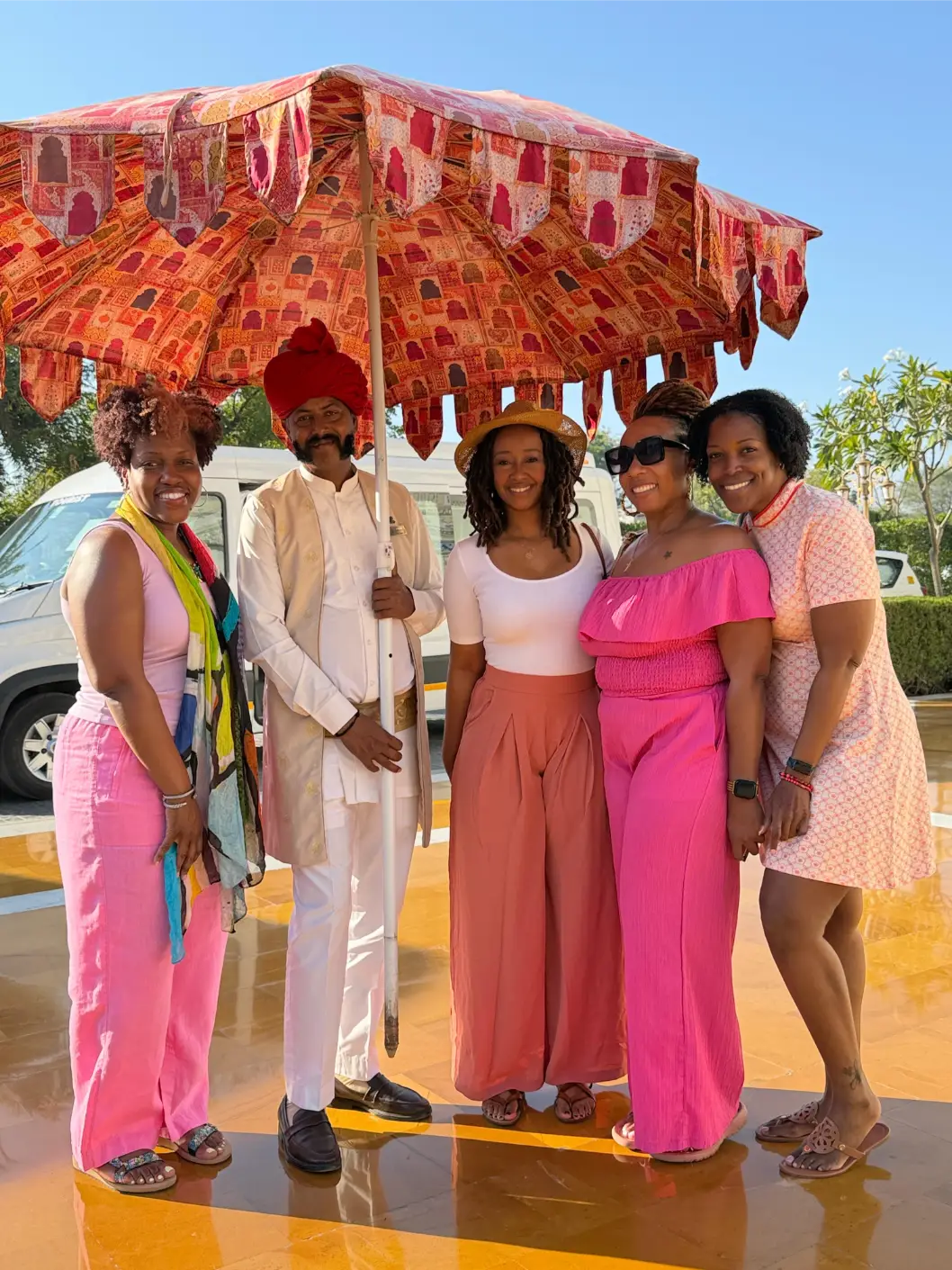 Five people smiling under a large patterned umbrella with a man dressed in white and a red turban.