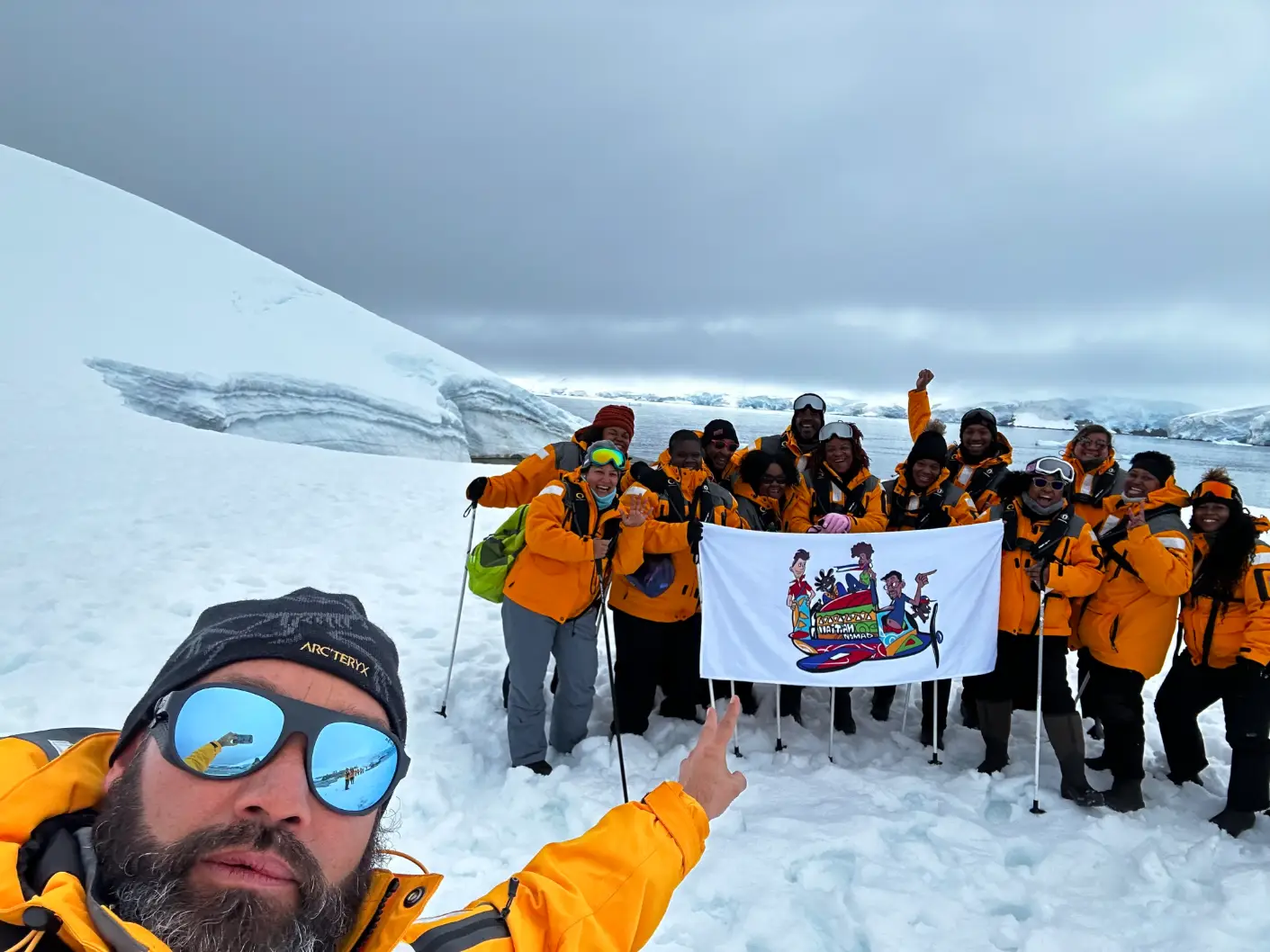Group in orange jackets on snowy terrain holding a white flag with colorful cartoon figures.