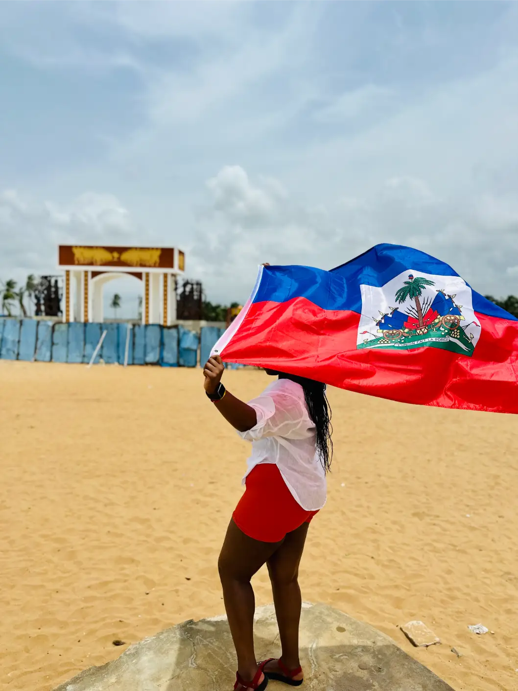 Person holding a Haitian flag on a sandy beach with a cloudy sky and structure in the background.