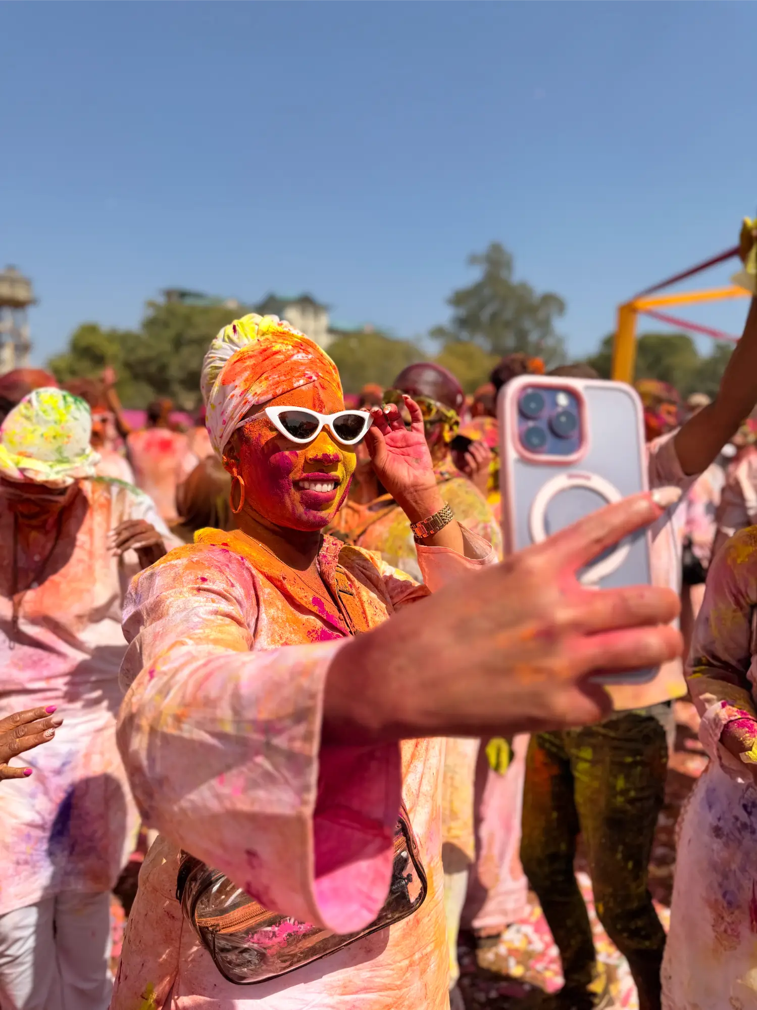 Woman covered in colorful powder wearing sunglasses takes a selfie at a vibrant festival crowd.