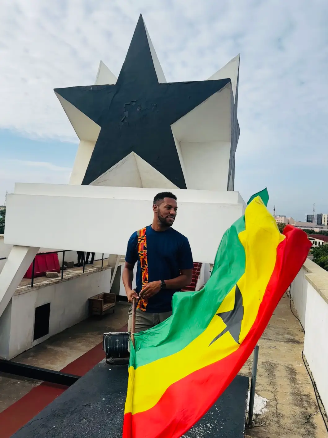 Man holding a Ghanaian flag waving in front of the Black Star Gate monument in Accra.