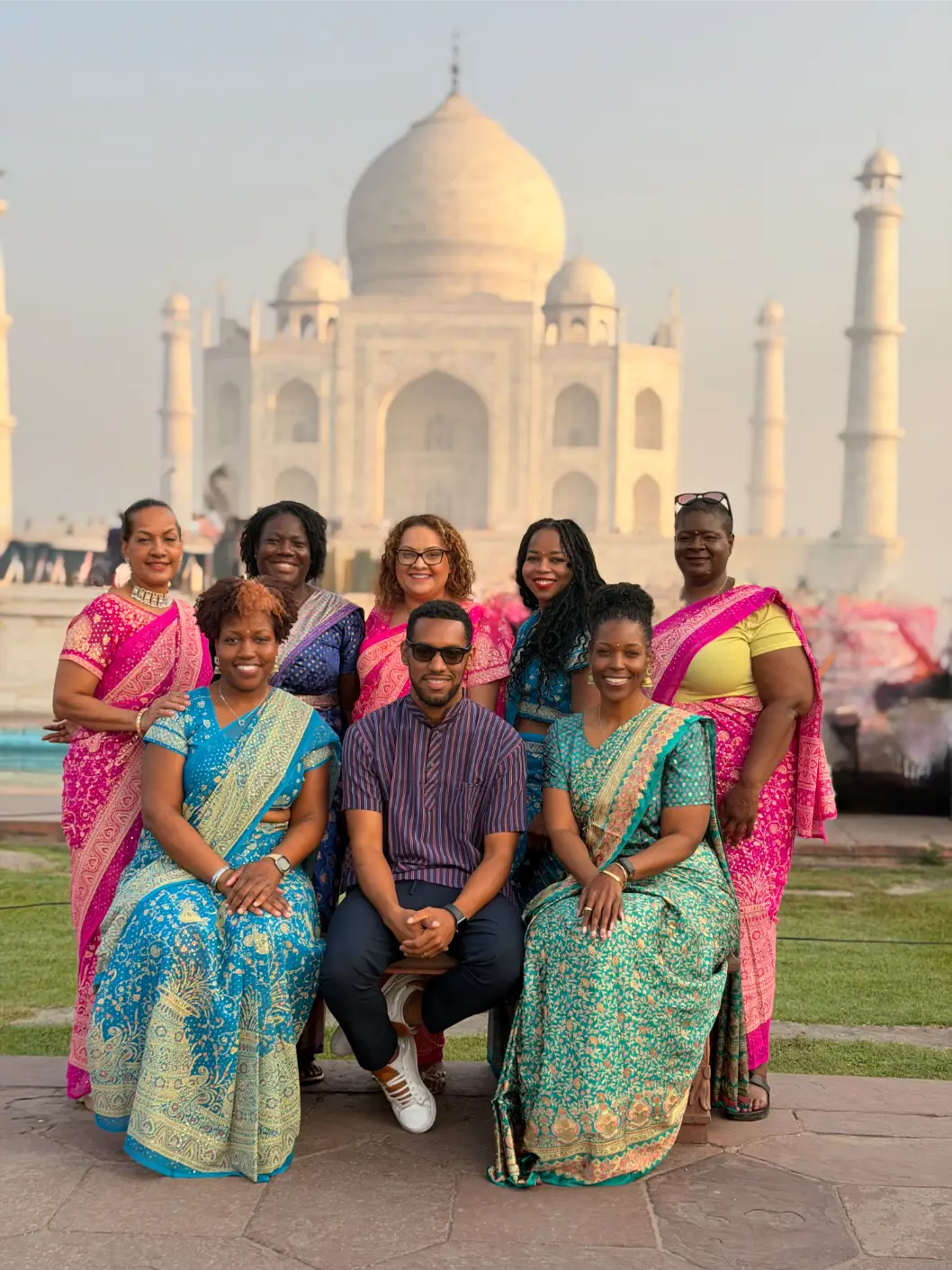 Group of eight people in colorful traditional clothing smiling in front of the Taj Mahal at sunset.