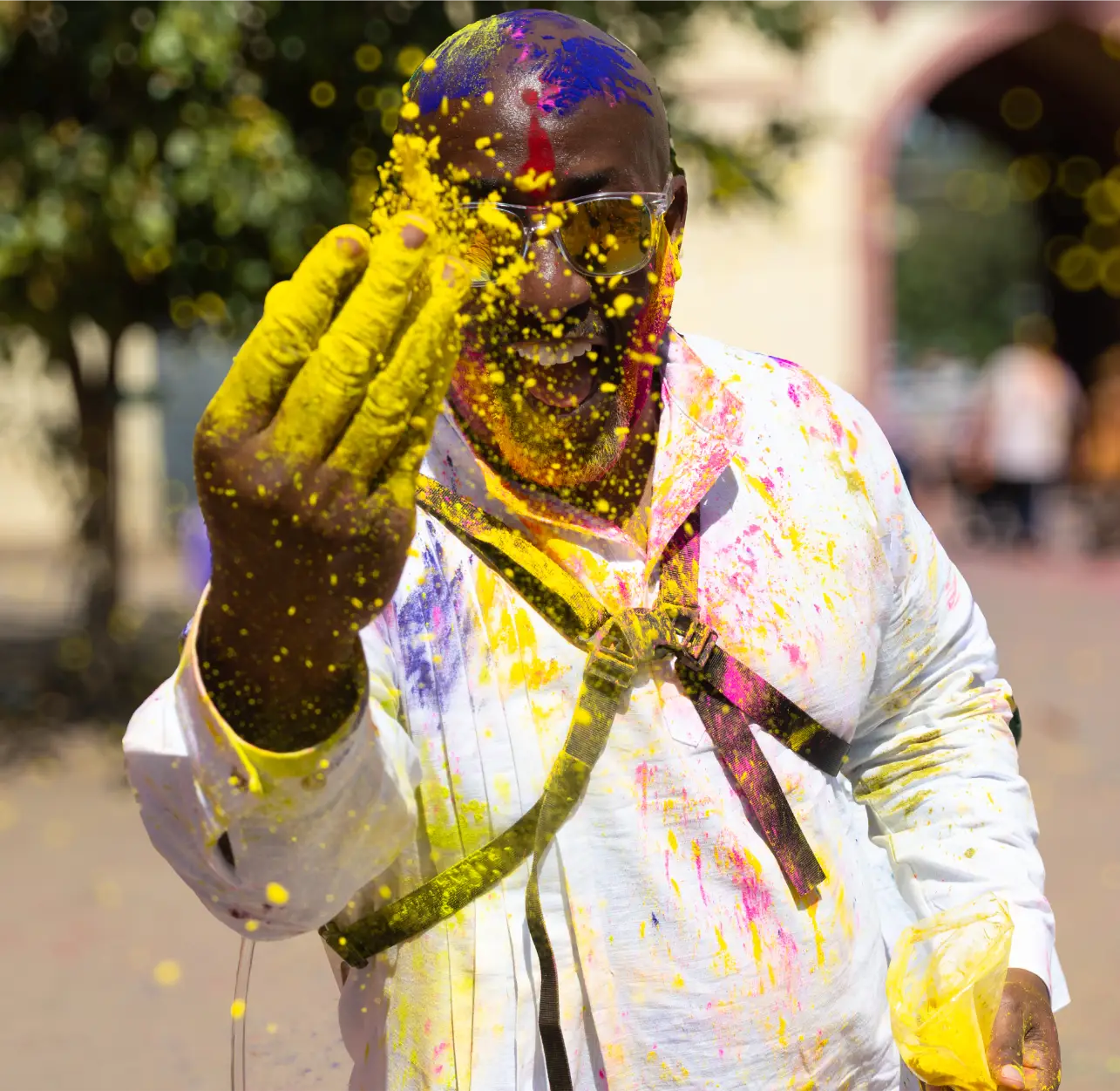 Man joyfully throwing yellow powder during Holi festival, covered in colorful powders wearing sunglasses.