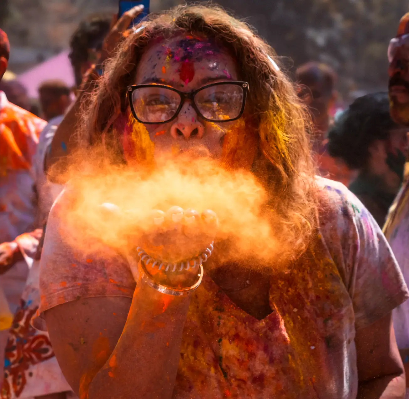 Person with glasses blowing orange powder during colorful Holi festival celebration.