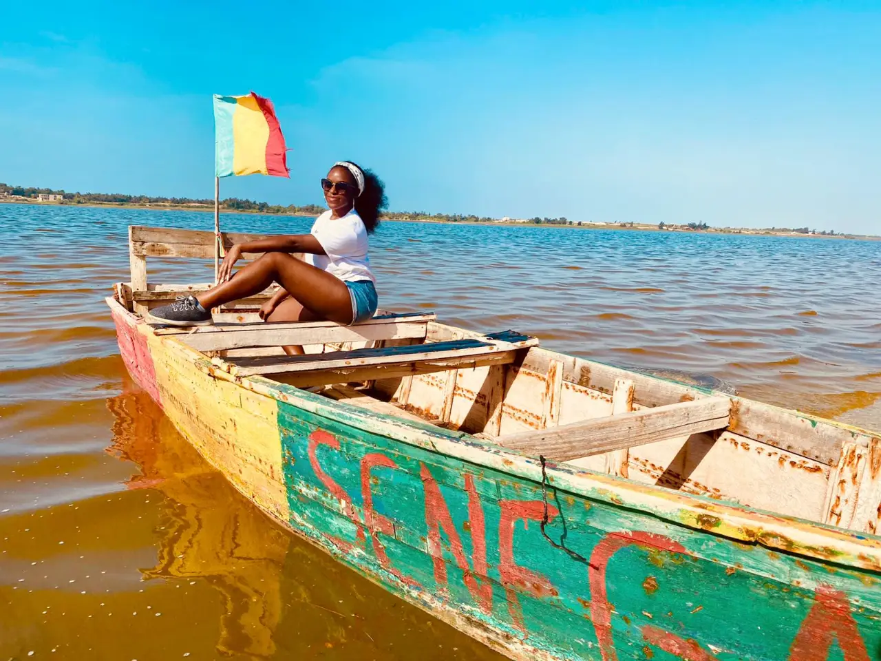 Woman in sunglasses and headband sitting on a colorful wooden boat on a calm lake.