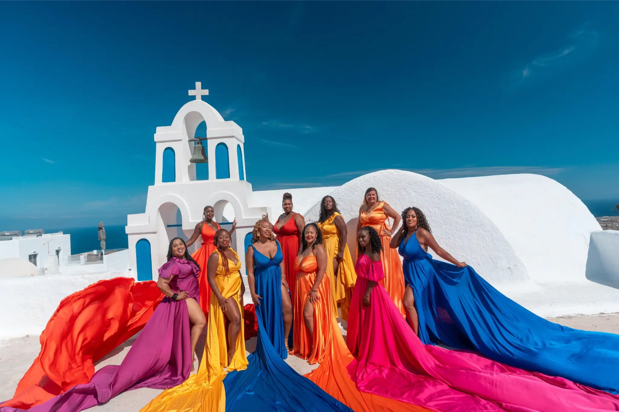 Group of women in vibrant colored dresses with long trains posing in front of a white church with a bell tower.