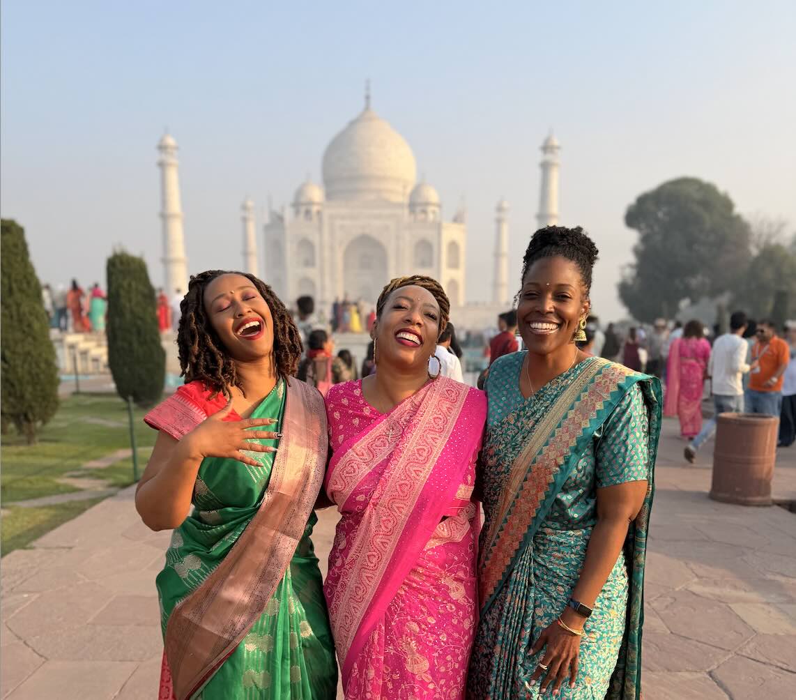 Three women in colorful saris laughing together in front of the Taj Mahal.