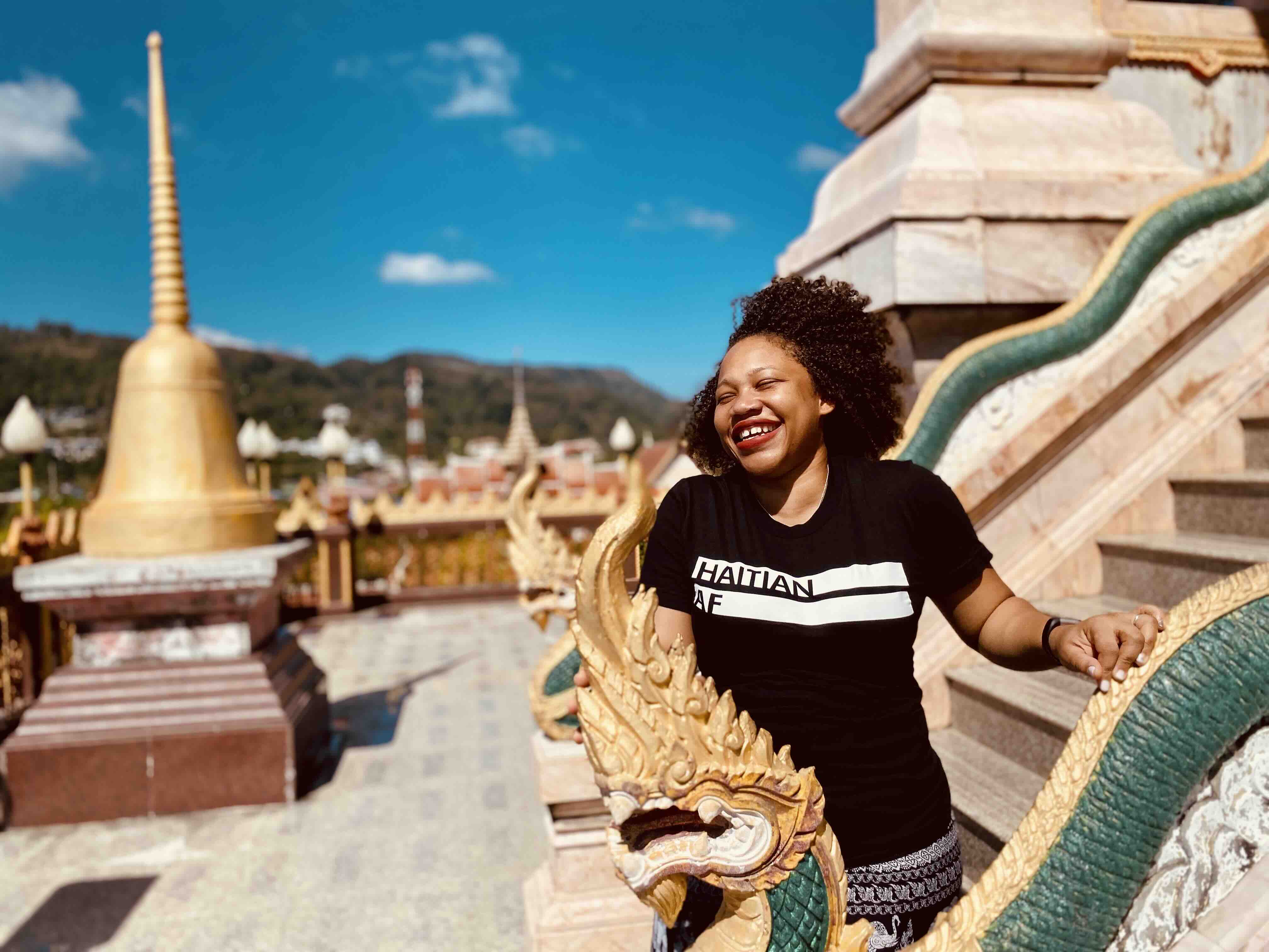 A traveler in a Haitian Nomad t-shirt smiling atop a golden temple staircase overlooking a stupa.