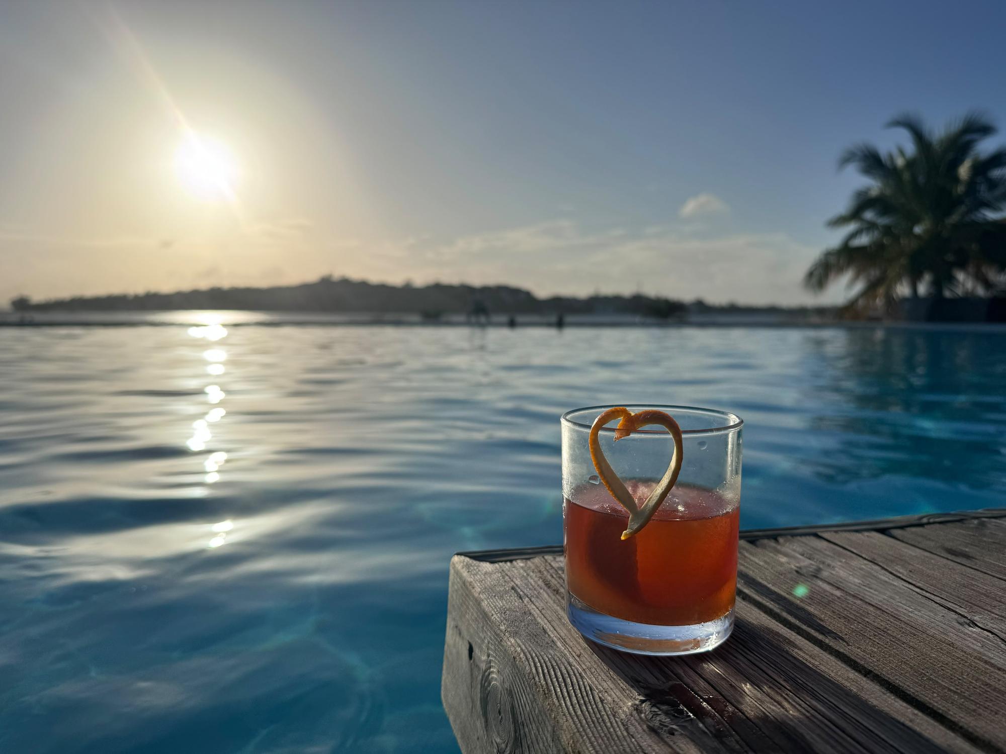 Cocktail garnished with a heart-shaped orange peel beside an infinity pool overlooking the ocean at sunset.