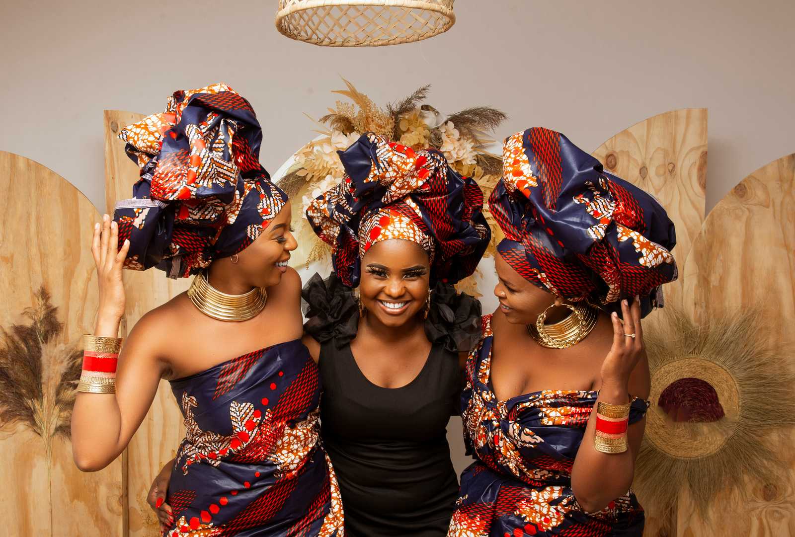 Three women smiling together in colorful African headwraps and gold jewelry.
