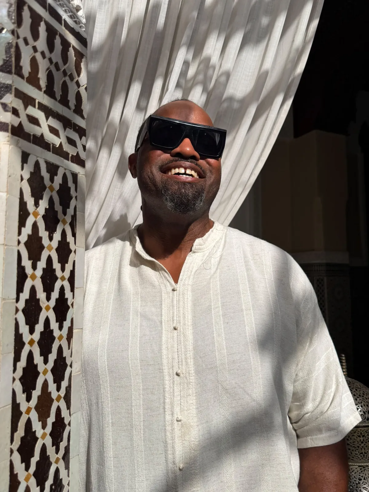 A smiling Haitian Nomad traveler in a white tunic beside a zellige-tiled column in Morocco