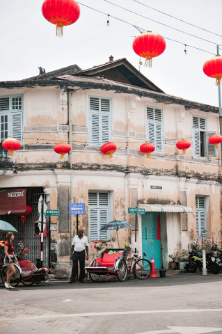  historic colonial-style building with weathered walls adorned with red Chinese lanterns, located at a street corner with rickshaws parked nearby and pedestrians enjoying the scene.