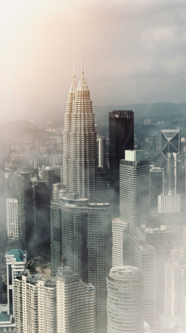 A misty aerial view of Kuala Lumpur's skyline, prominently featuring the Petronas Twin Towers surrounded by modern skyscrapers and soft sunlight filtering through the clouds.