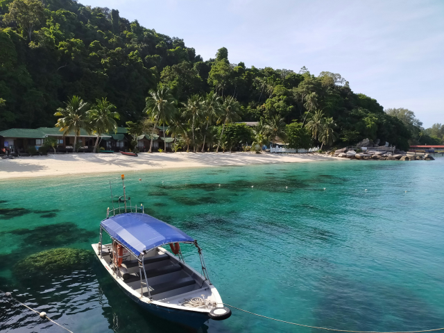 A picturesque tropical beach on an island with clear turquoise water, a small boat anchored in the foreground, and lush greenery and palm trees surrounding the sandy shore.