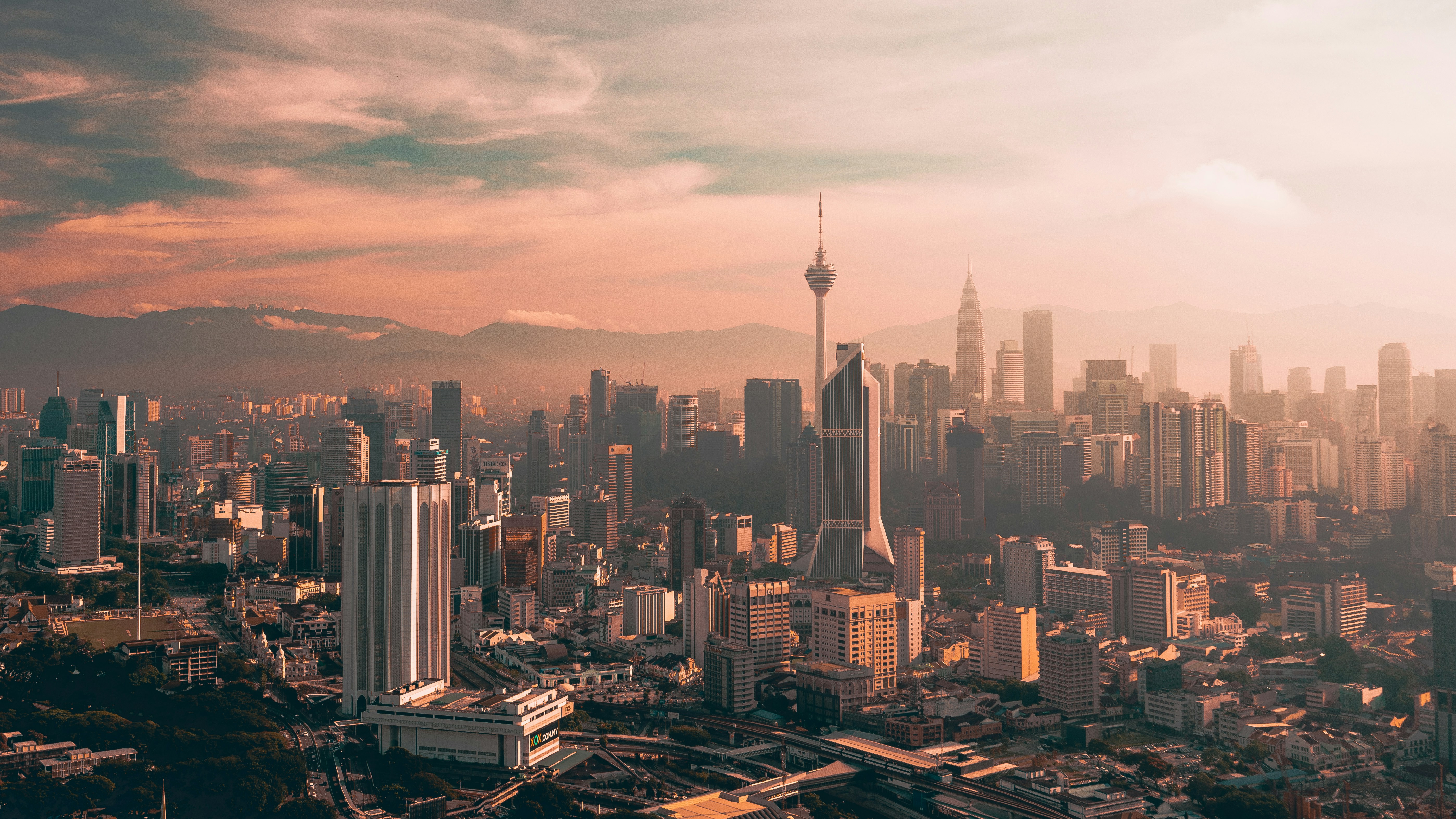 Aerial view of Kuala Lumpur's skyline during sunset, featuring prominent landmarks like the Kuala Lumpur Tower and Petronas Twin Towers, with the cityscape framed by distant mountains.