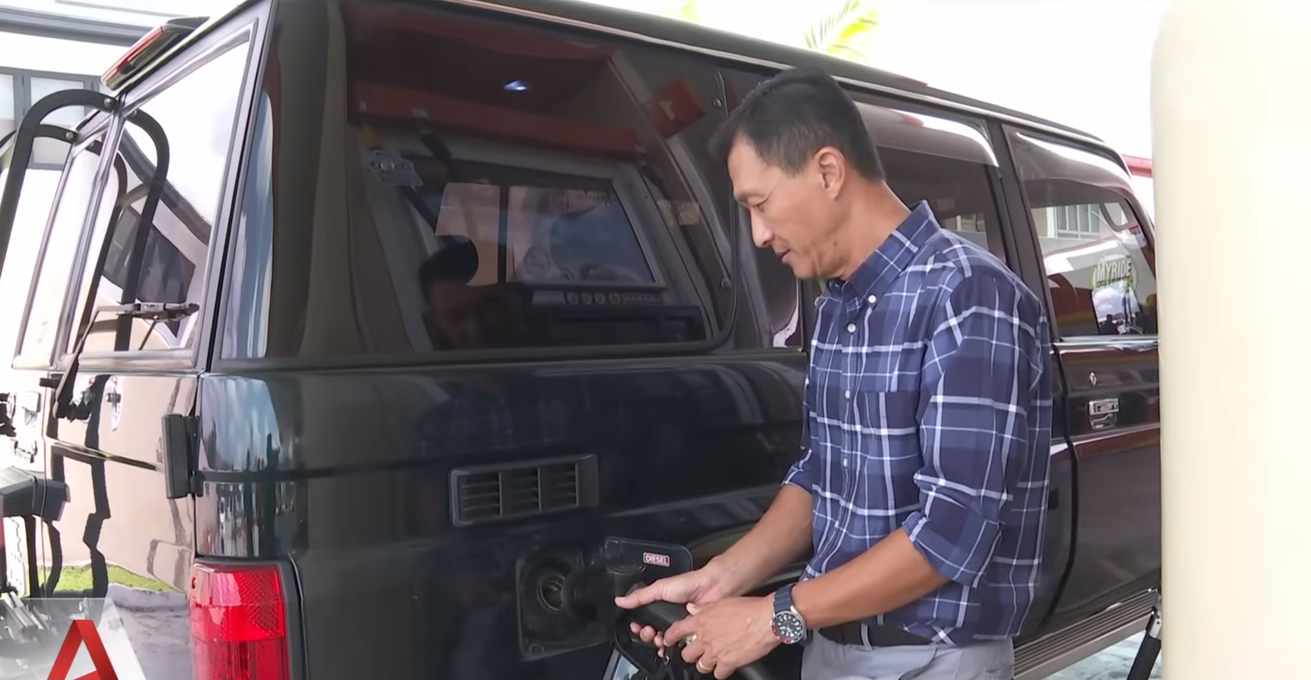 Man refueling a dark green SUV at a gas station, wearing a blue plaid shirt. The reflection of the gas station and surroundings is visible on the vehicle's windows.