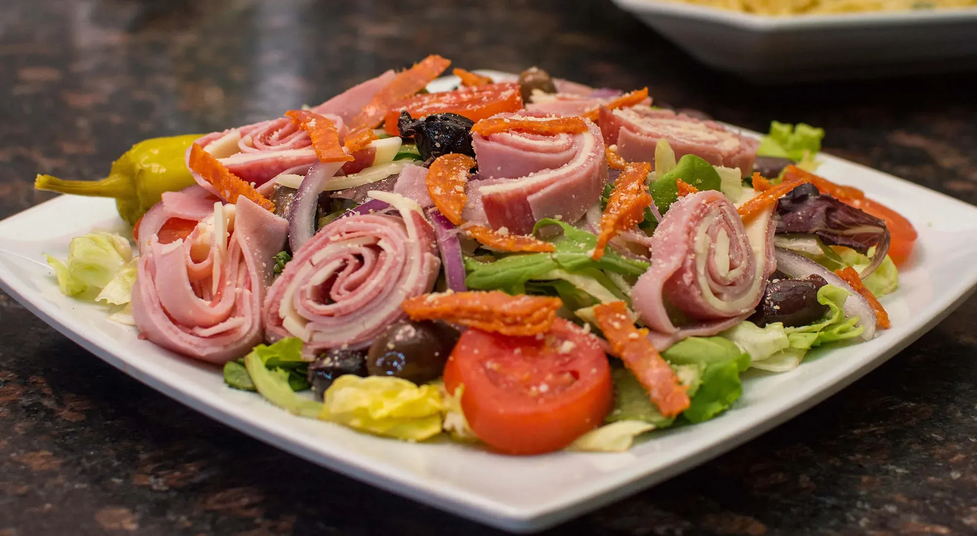 Plated salad with lettuce, tomato, rolled deli meats, olives, shredded cheese, and a pepperoncini pepper.