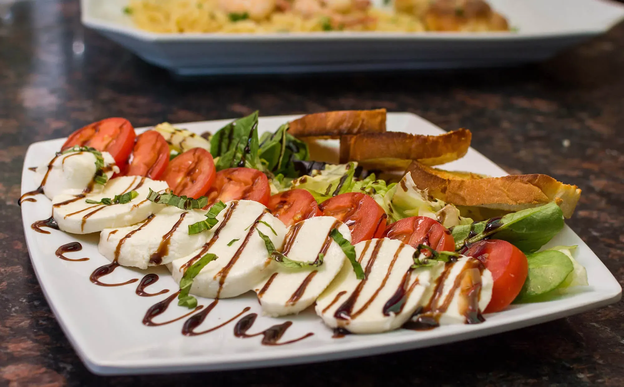 Caprese salad with tomato, mozzarella, basil, balsamic glaze, and toasted bread on a white plate.