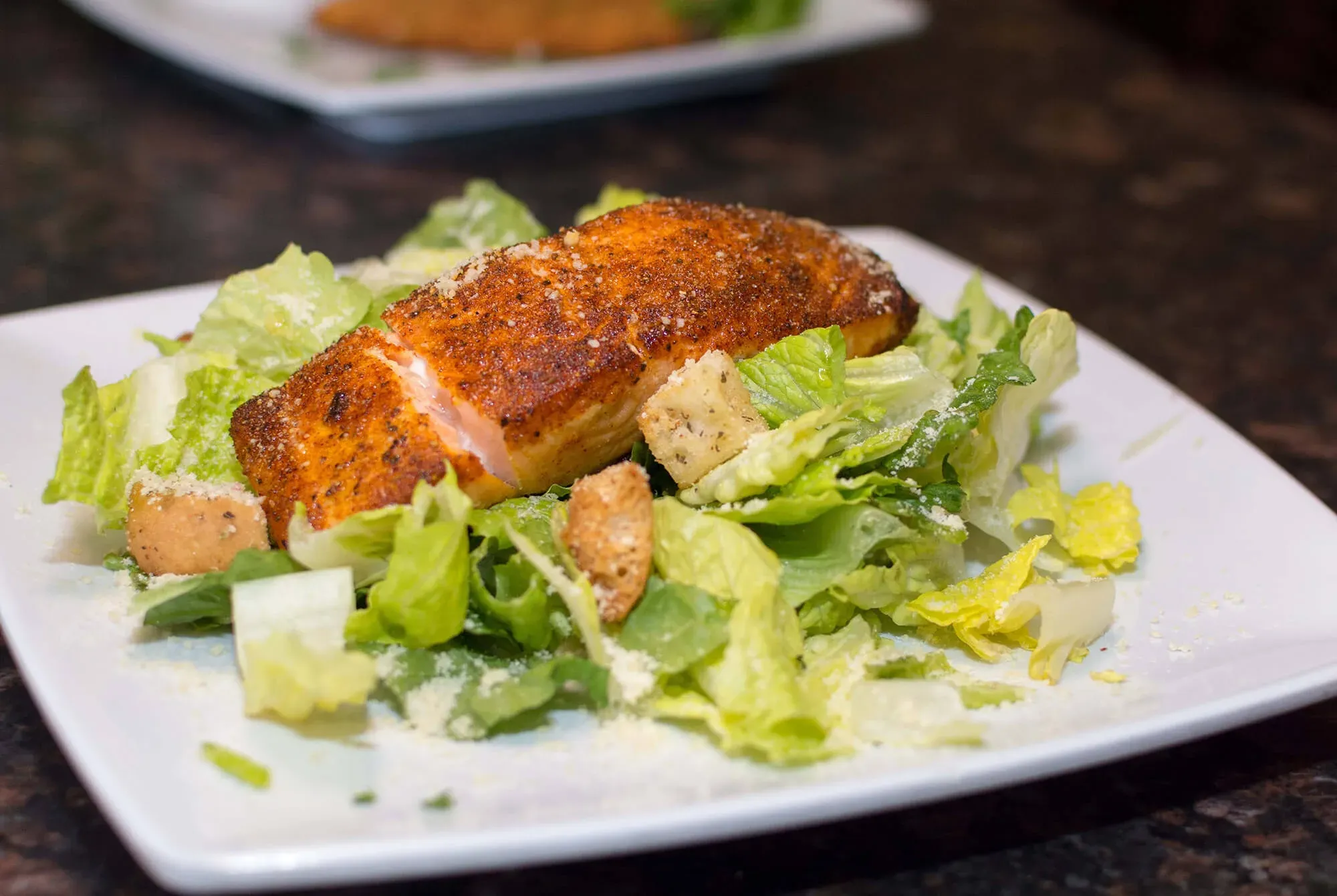 Blackened salmon fillet on romaine lettuce salad with croutons and grated cheese on white plate.