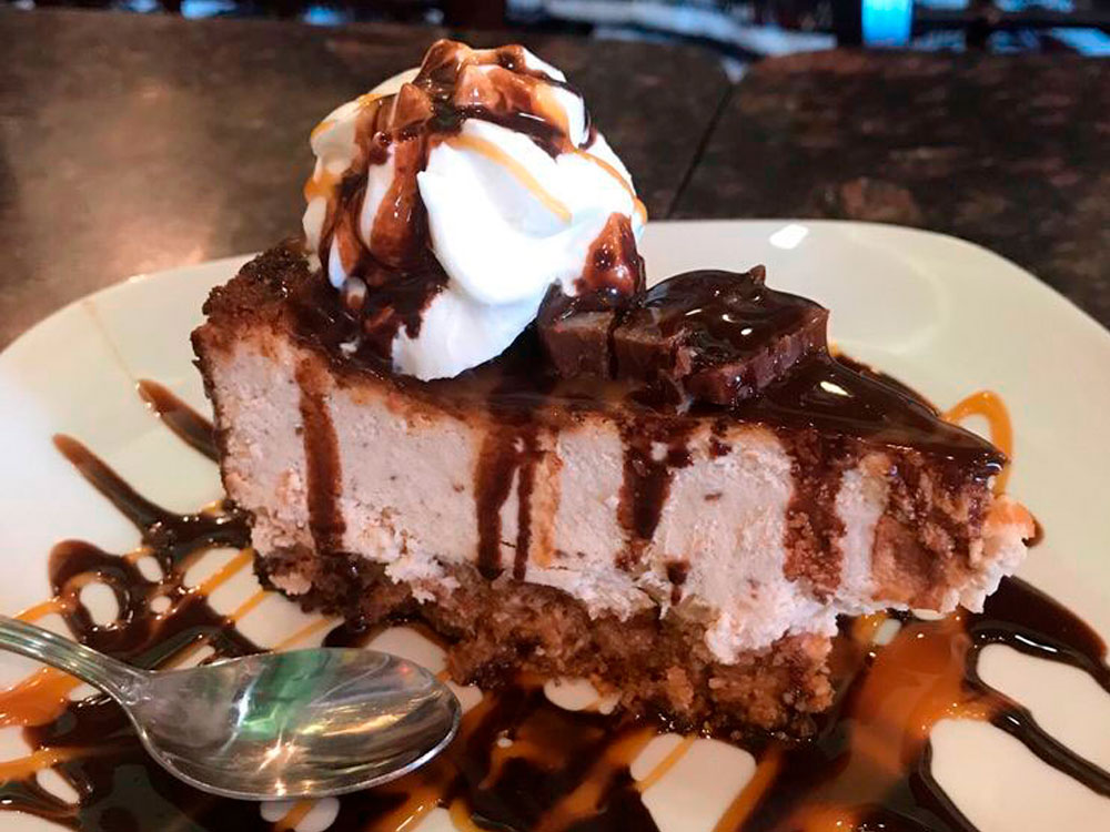 Slice of cheesecake topped with whipped cream, chocolate drizzle, and a small brownie square on a white plate.