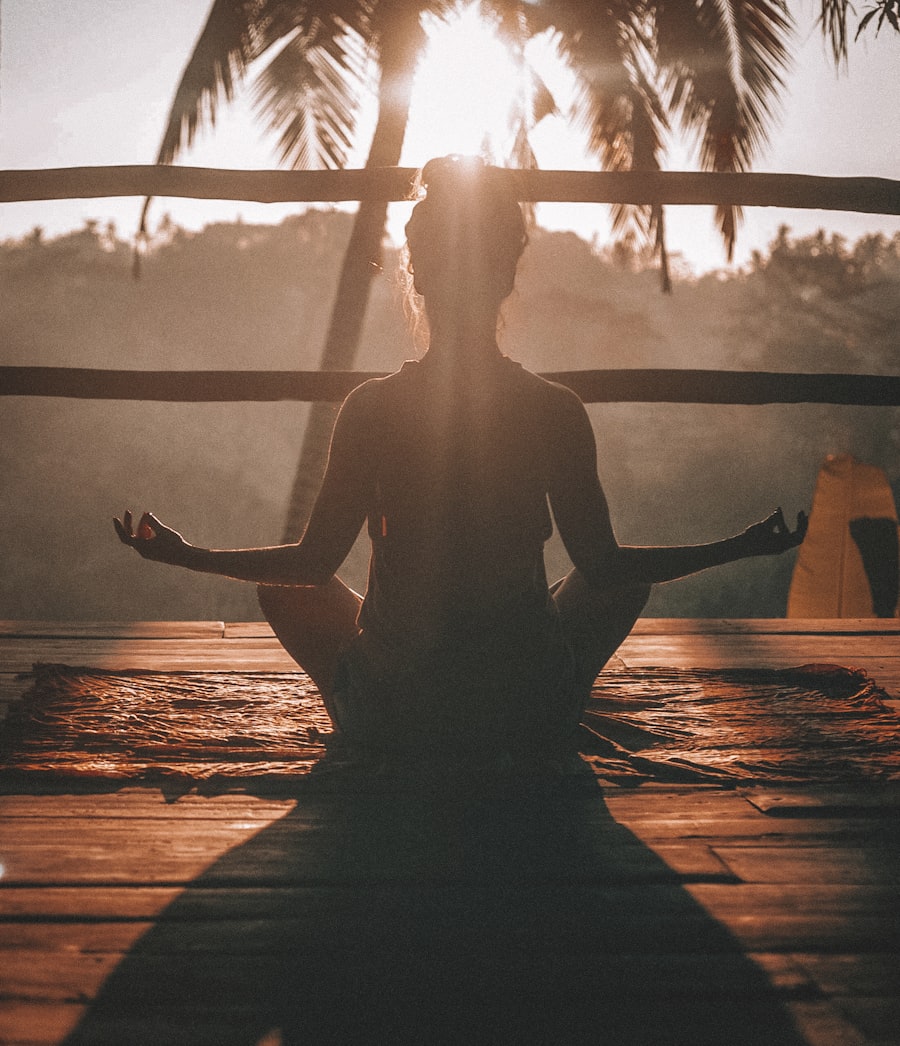 Person sitting cross-legged meditating on a mat under palm trees at sunset.
