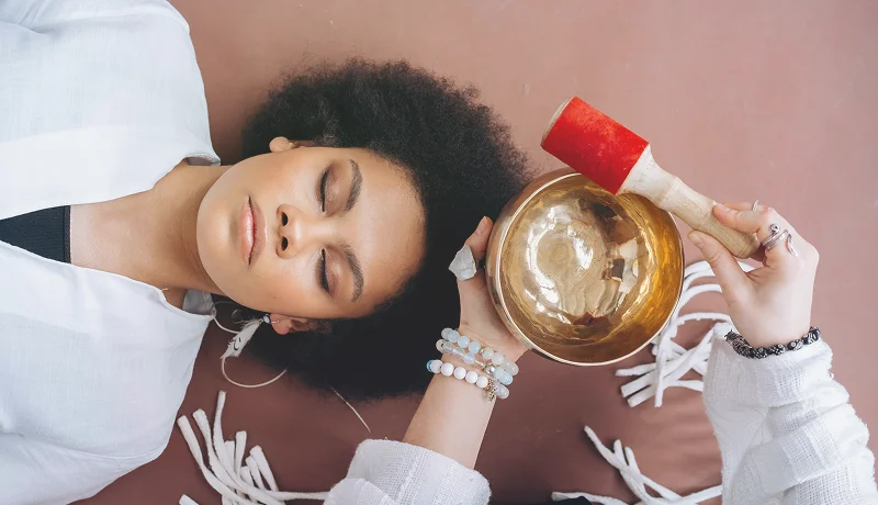Woman lying down with eyes closed, holding a golden singing bowl and mallet near her head.