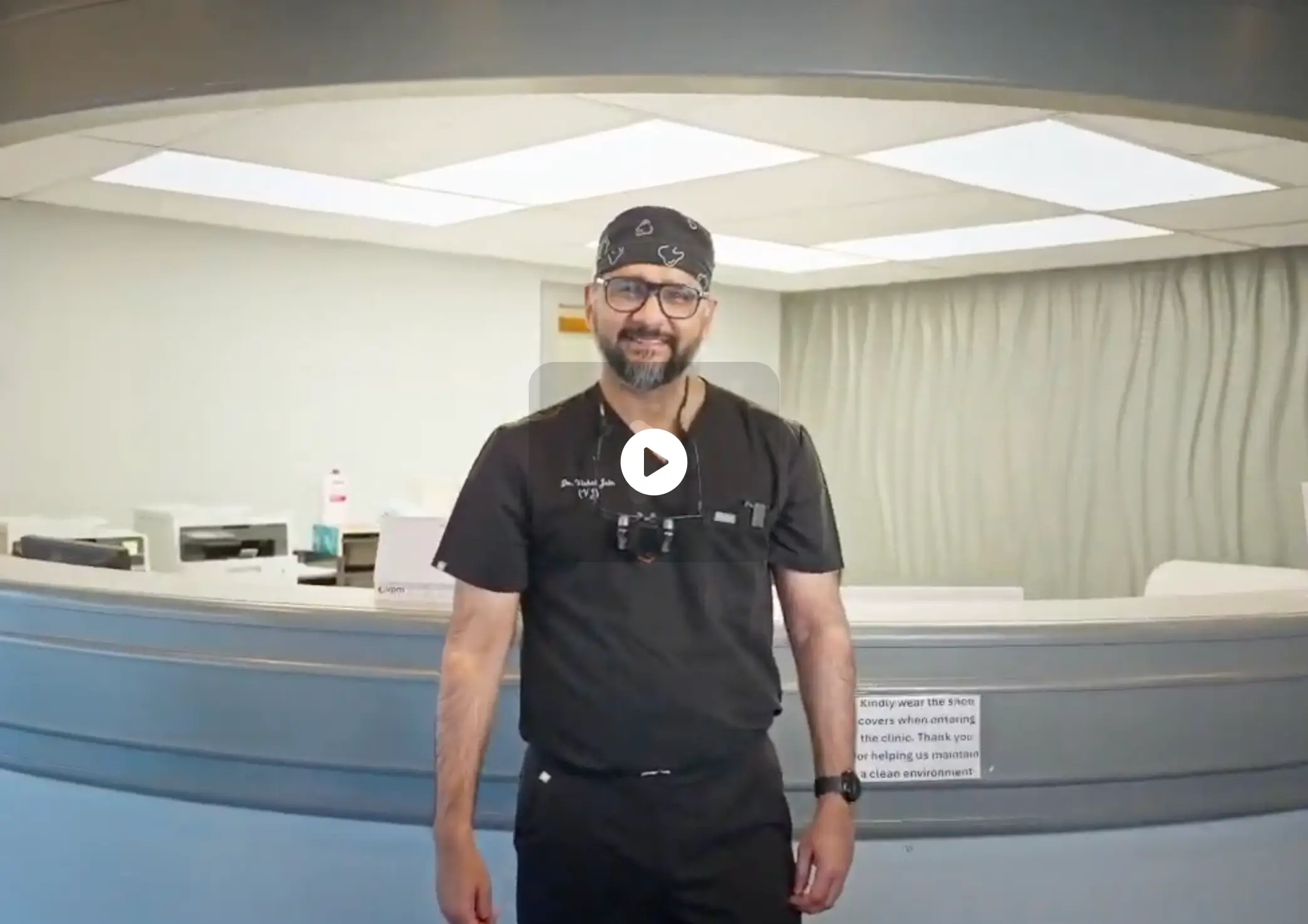 Smiling male dentist in black scrubs and glasses stands in a bright dental clinic reception area. La Perle Dental Center in Edmonton, AB TST 3T9