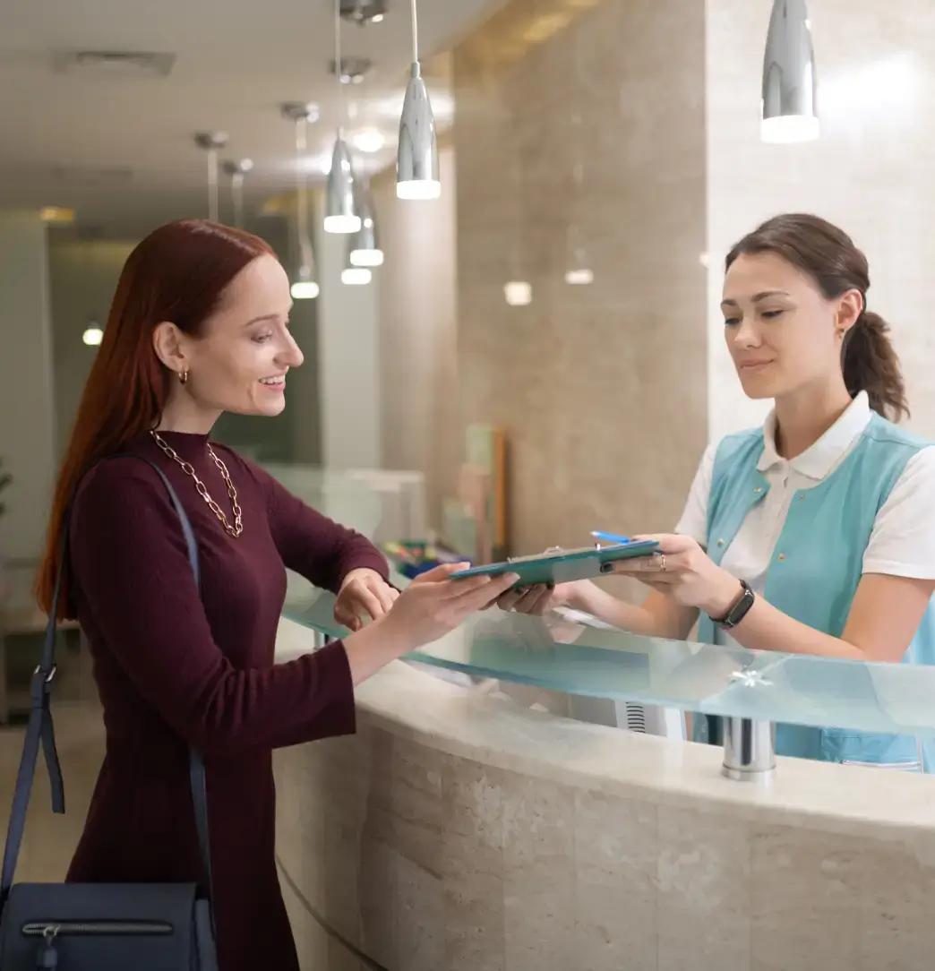 Woman at reception desk handing a clipboard to a healthcare worker in a mint vest under hanging lights. La Perle Dental Center in Edmonton, AB TST 3T9