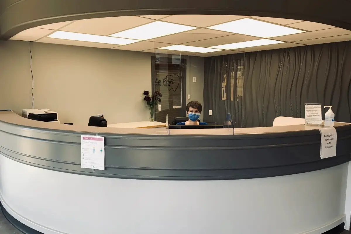 Reception desk with a masked person behind a clear shield and sanitizer on the counter. La Perle Dental Center in Edmonton, AB TST 3T9