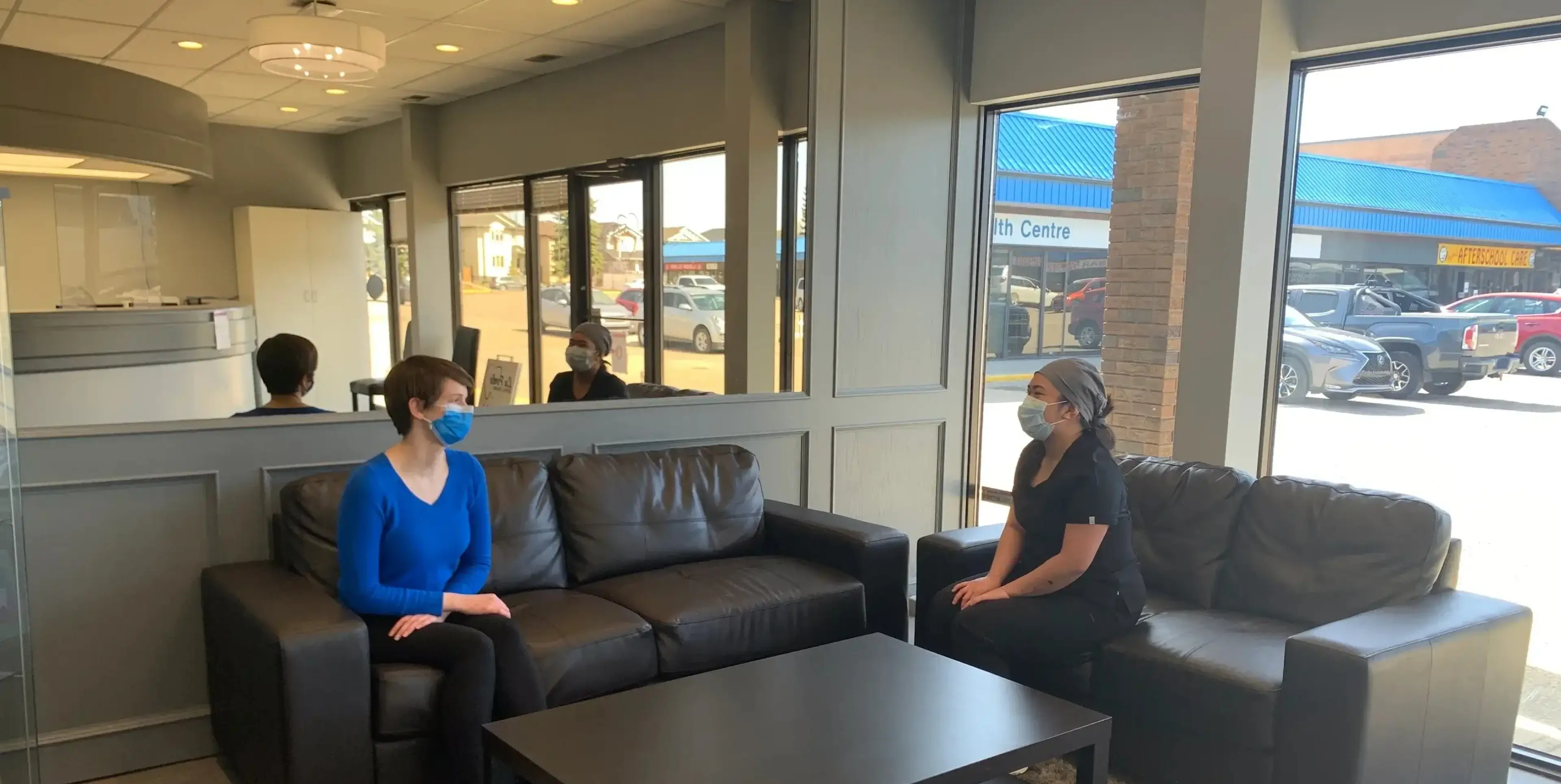Two masked women sitting on black sofas in a lobby with large windows showing parked cars outside. La Perle Dental Center in Edmonton, AB TST 3T9