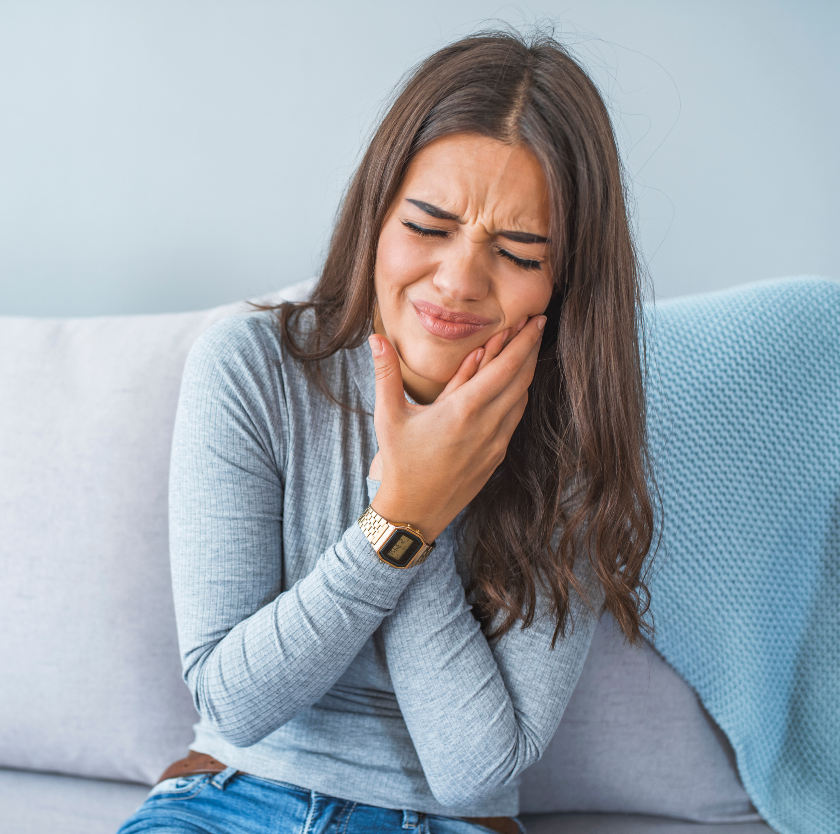 Young woman sitting on couch holding her jaw with a painful expression indicating a toothache. La Perle Dental Center in Edmonton, AB TST 3T9