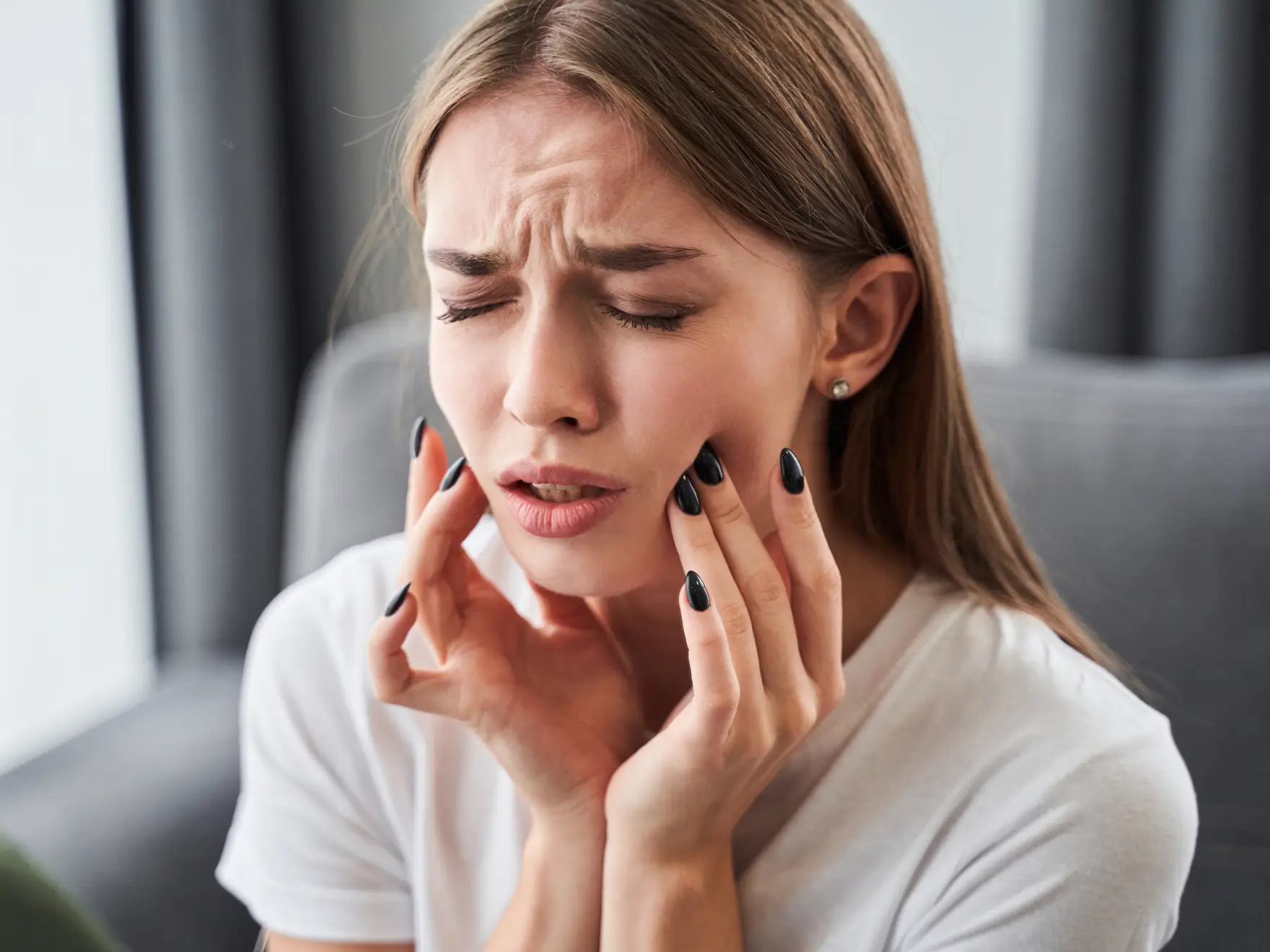 Woman wearing white shirt holding her jaw with both hands, showing pain or discomfort. La Perle Dental Center in Edmonton, AB TST 3T9