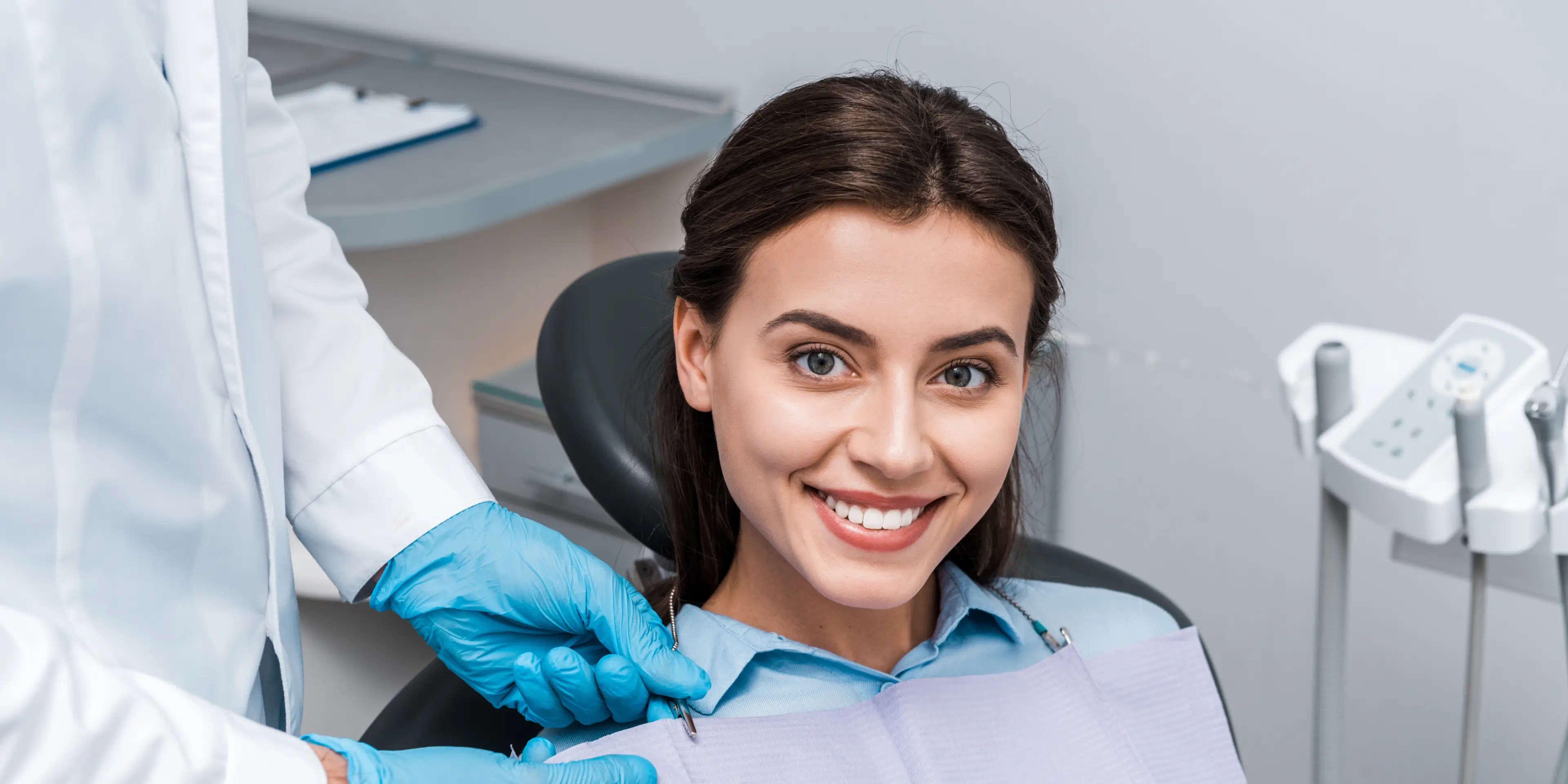 Smiling woman in dental chair with dentist attaching a protective bib on her. La Perle Dental Center in Edmonton, AB TST 3T9