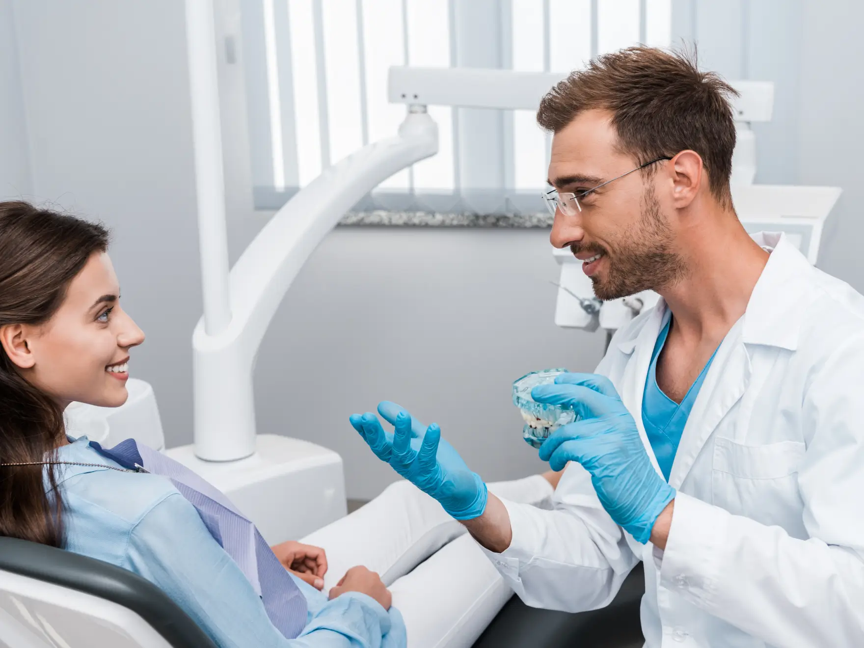 Dentist wearing gloves explaining a dental model to a smiling female patient in a dental office. La Perle Dental Center in Edmonton, AB TST 3T9