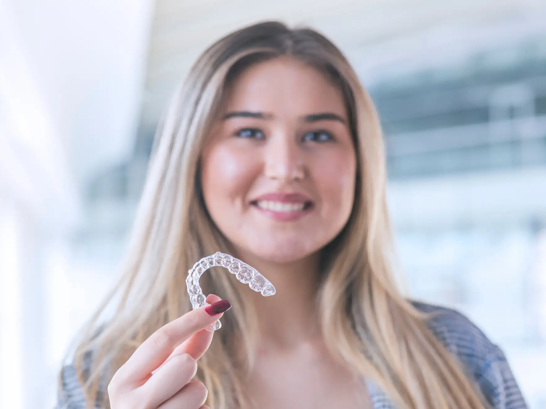Smiling woman holding a clear dental aligner between her fingers in a bright modern space. La Perle Dental Center in Edmonton, AB TST 3T9