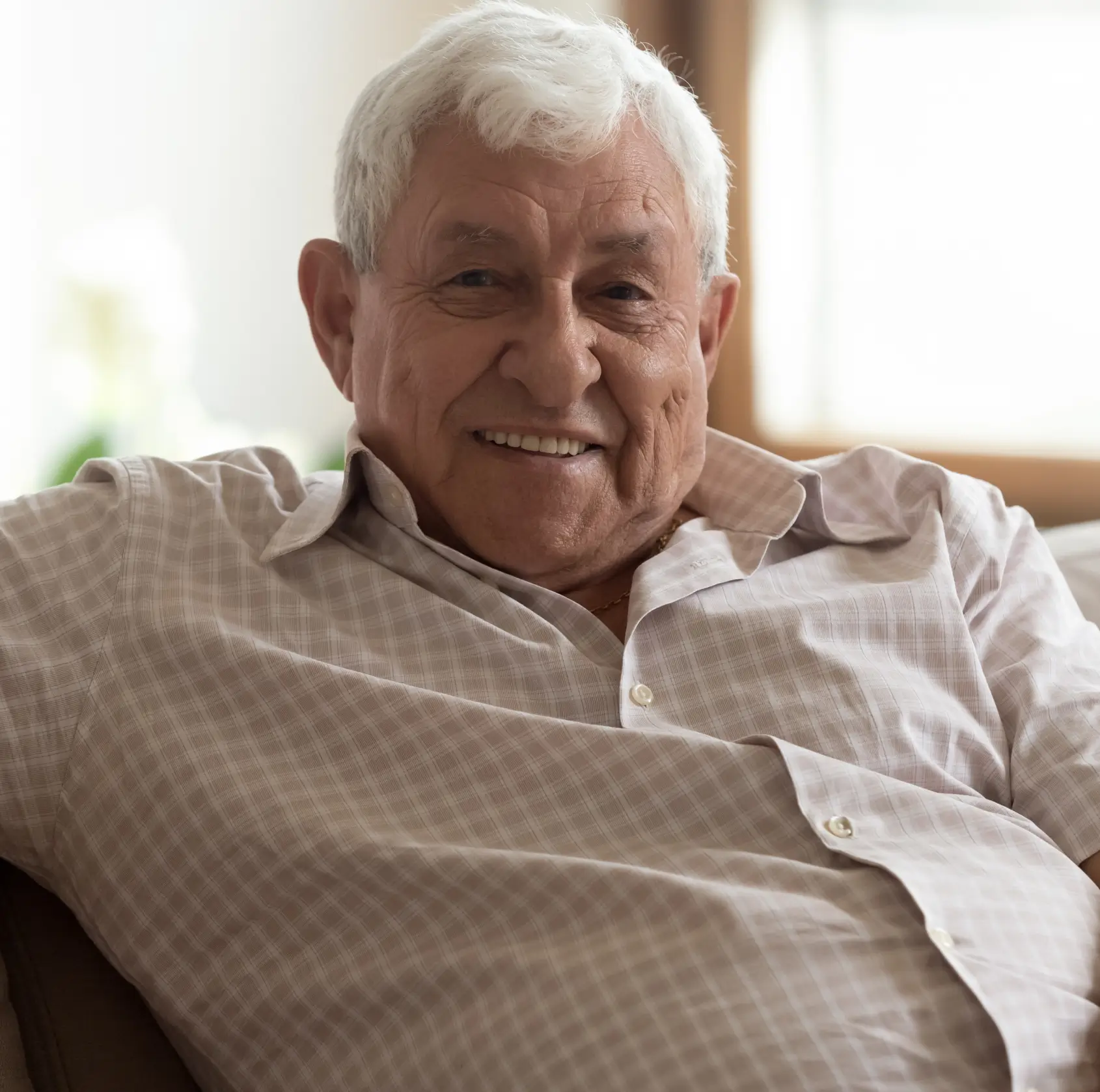 Smiling elderly man with white hair wearing a checkered shirt sitting indoors. La Perle Dental Center in Edmonton, AB TST 3T9