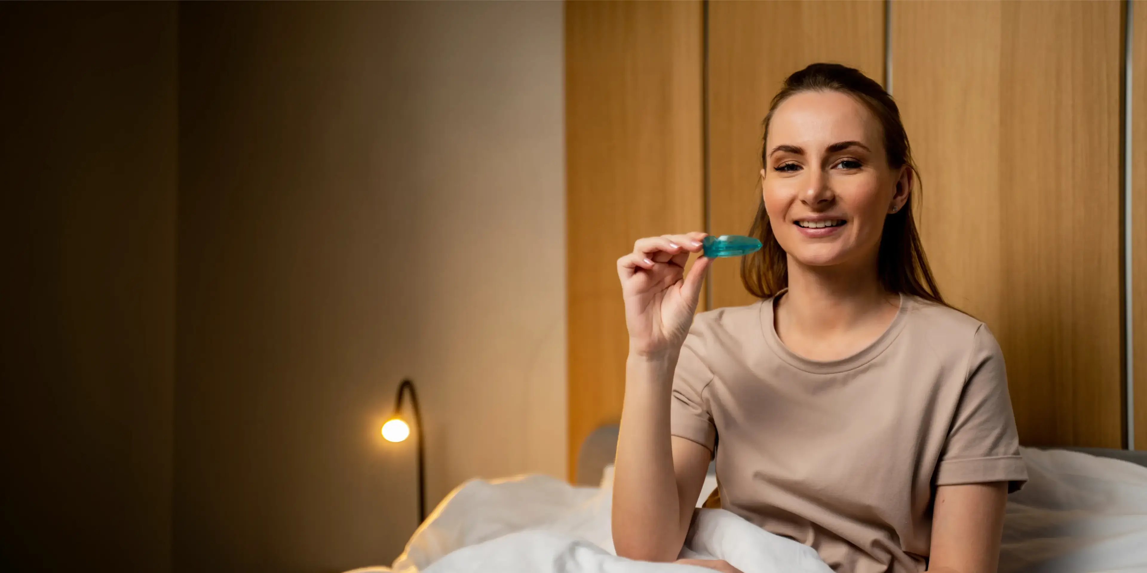 Smiling woman sitting on bed holding a blue night guard in her hand indoors. La Perle Dental Center in Edmonton, AB TST 3T9