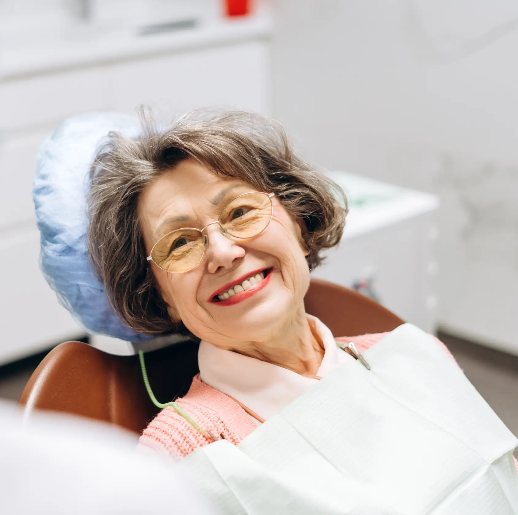 Smiling elderly woman with glasses sitting in a dental chair wearing a dental bib. La Perle Dental Center in Edmonton, AB TST 3T9