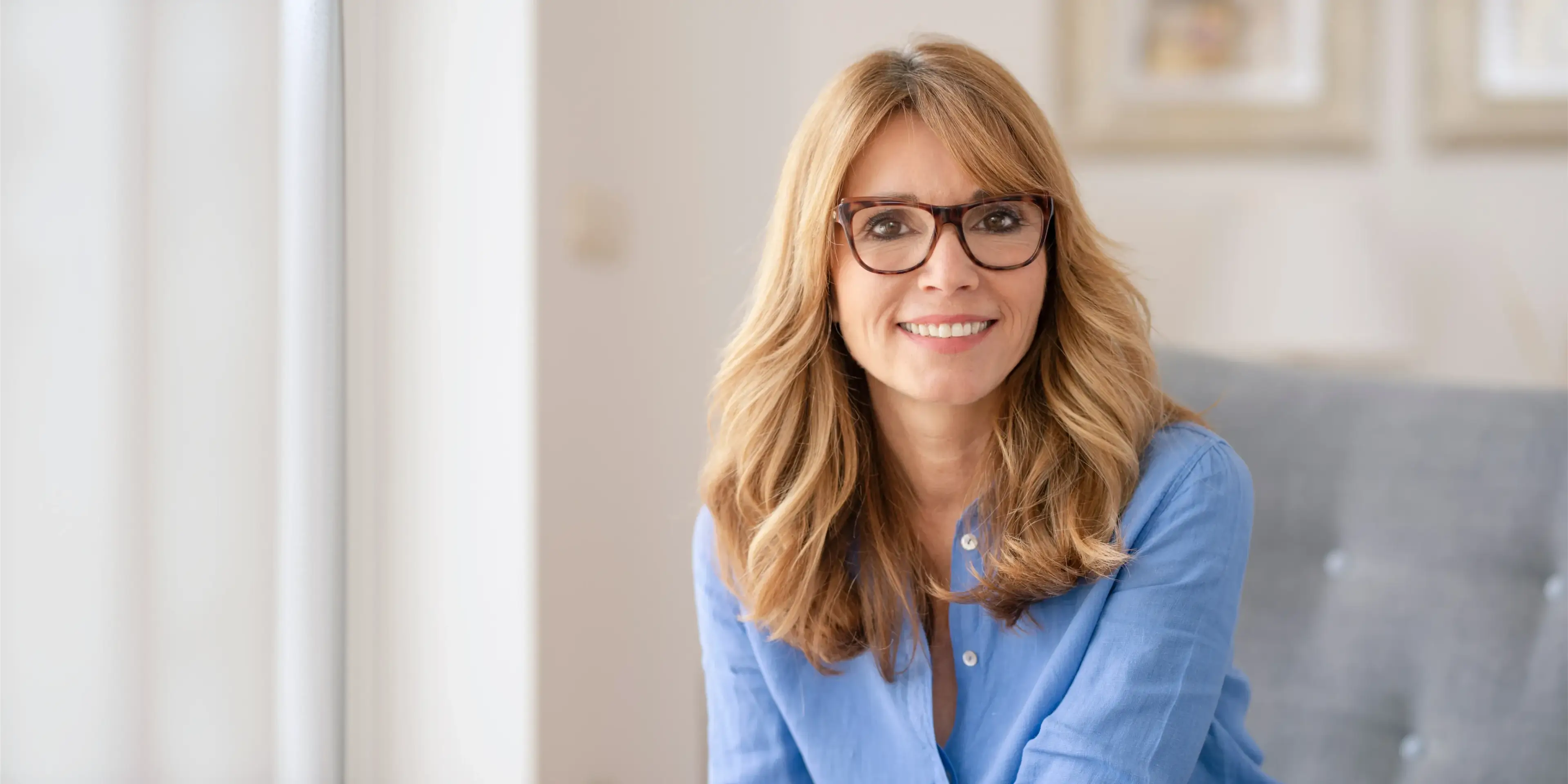 Smiling woman with blonde hair and glasses wearing a blue shirt, sitting indoors on a gray couch. La Perle Dental Center in Edmonton, AB TST 3T9