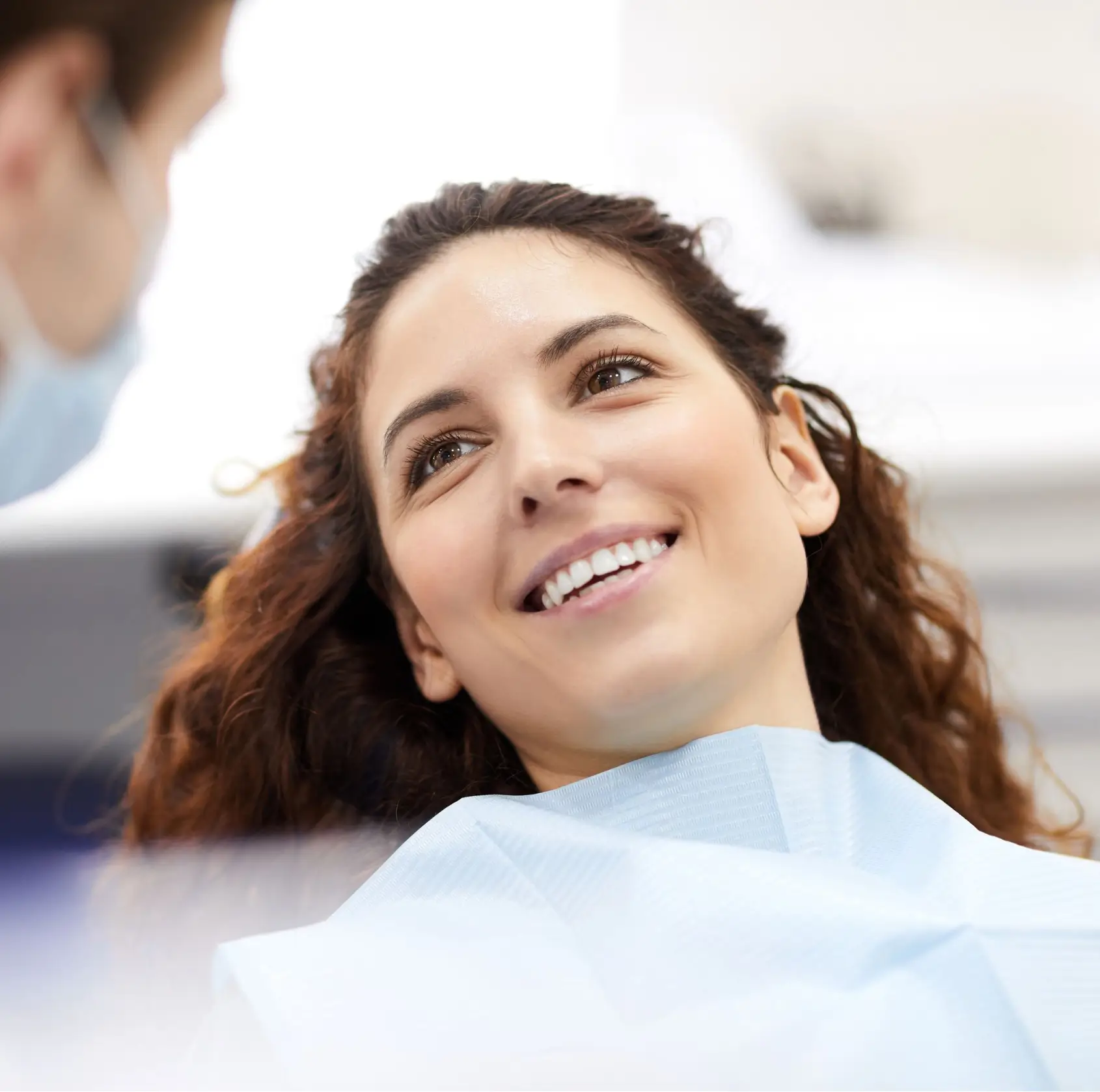 Smiling woman with curly hair sitting in a dental chair wearing a protective bib. La Perle Dental Center in Edmonton, AB TST 3T9