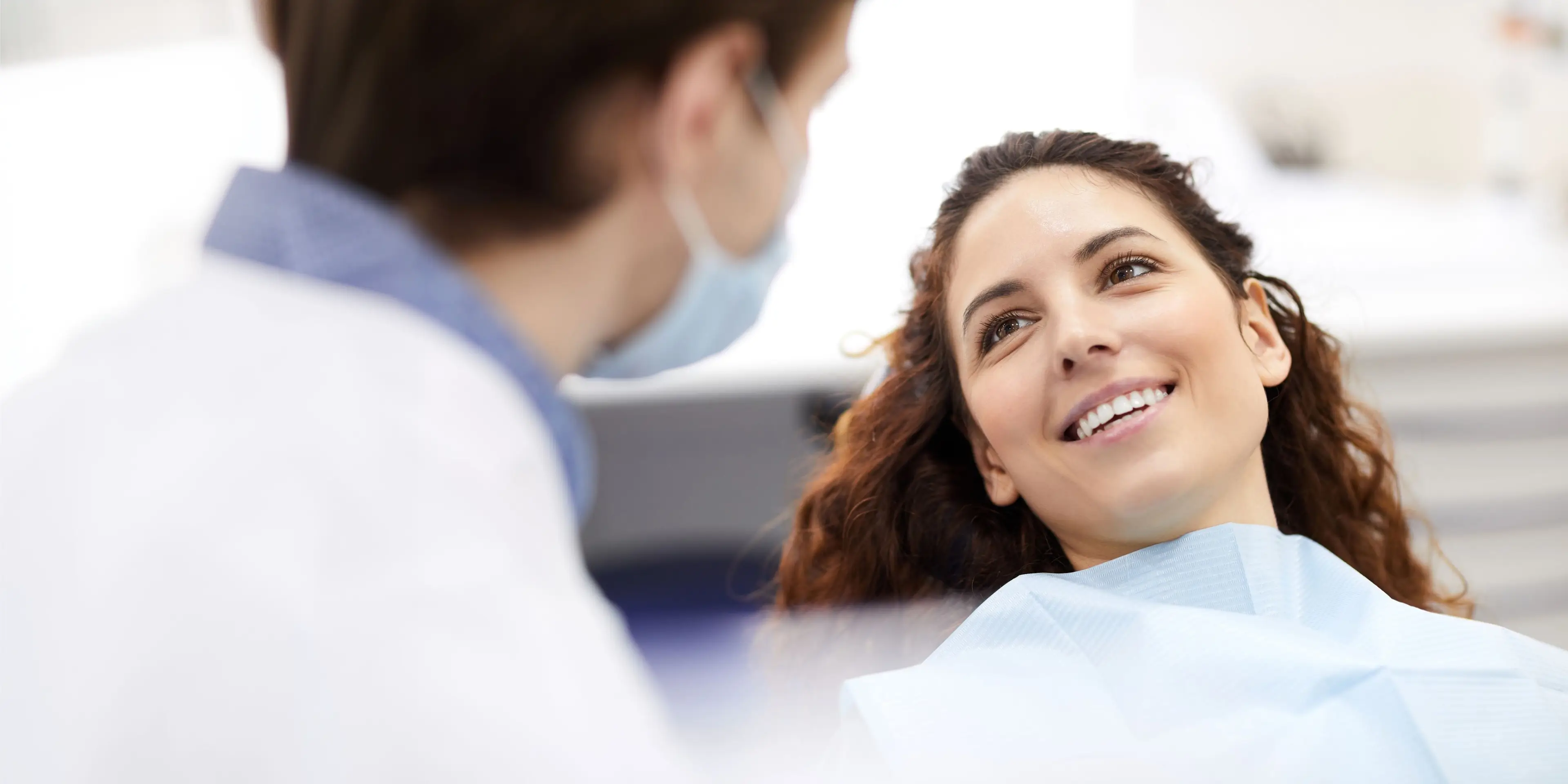 Smiling woman in dental chair looking at masked dentist in a clinic setting. La Perle Dental Center in Edmonton, AB TST 3T9