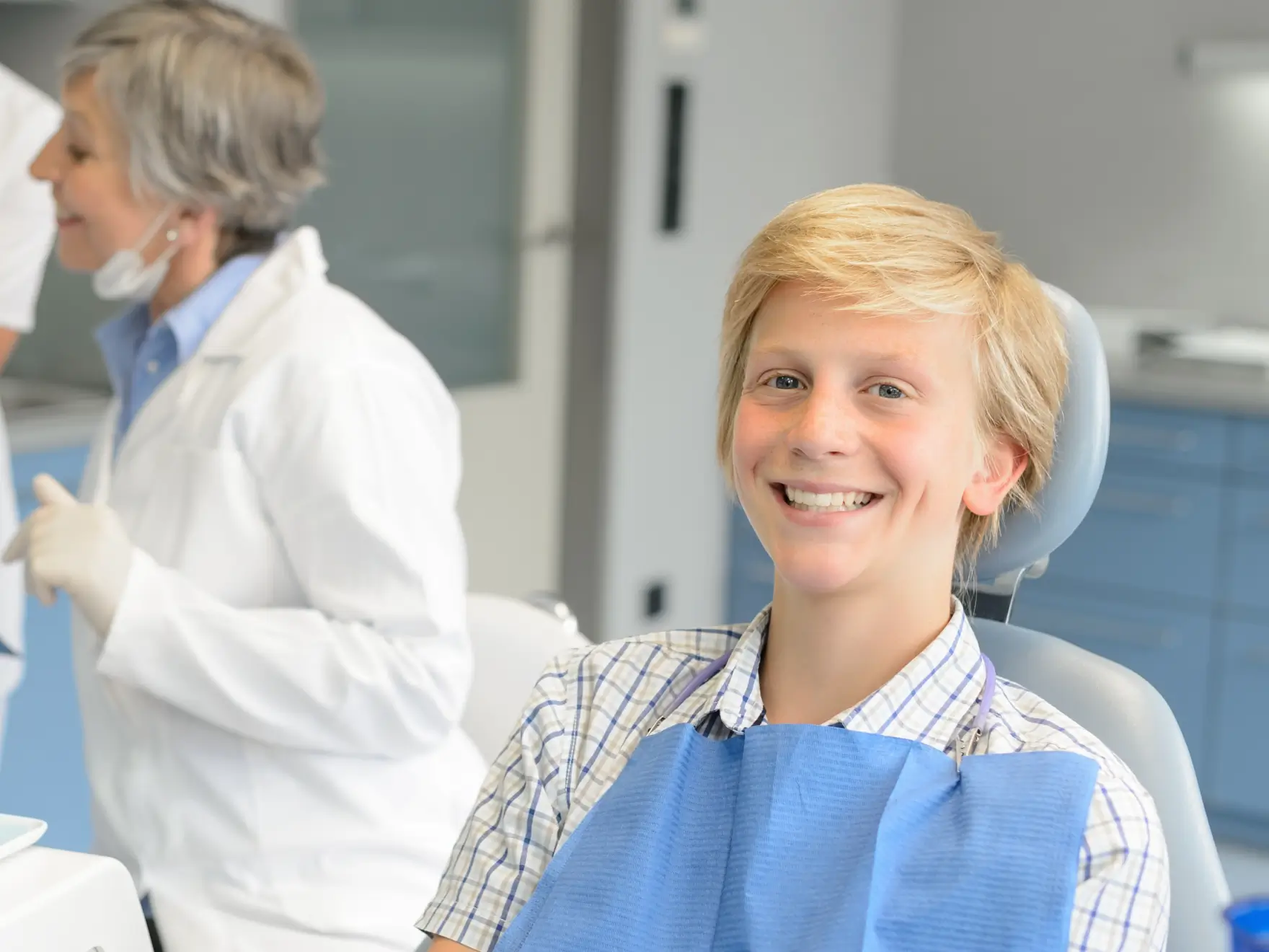 Smiling boy sitting in a dental chair with a blue bib, and a female dentist in the background. La Perle Dental Center in Edmonton, AB TST 3T9