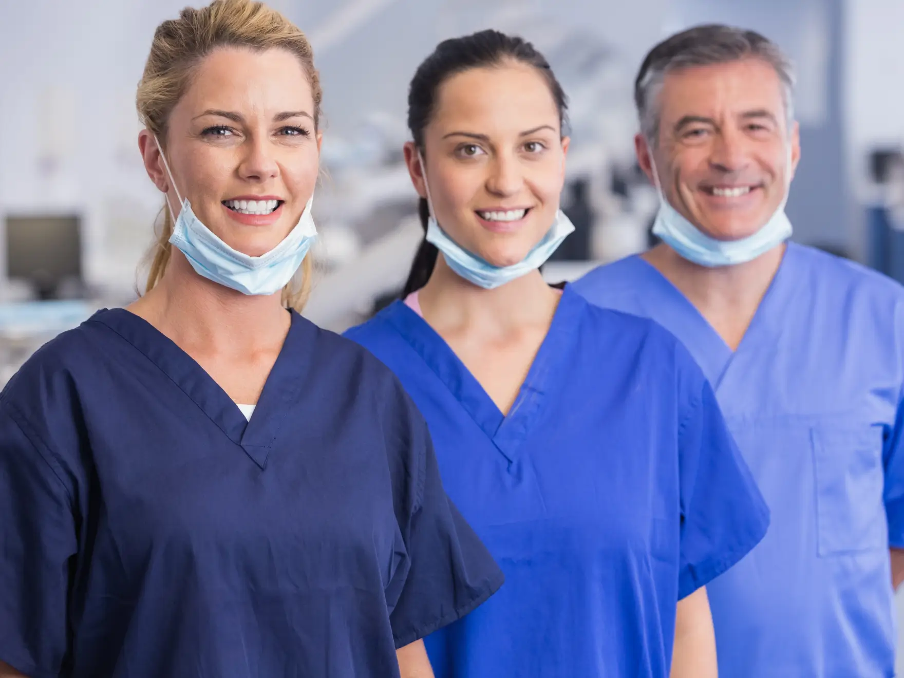 Three smiling healthcare workers in scrubs with face masks pulled down around their necks. La Perle Dental Center in Edmonton, AB TST 3T9