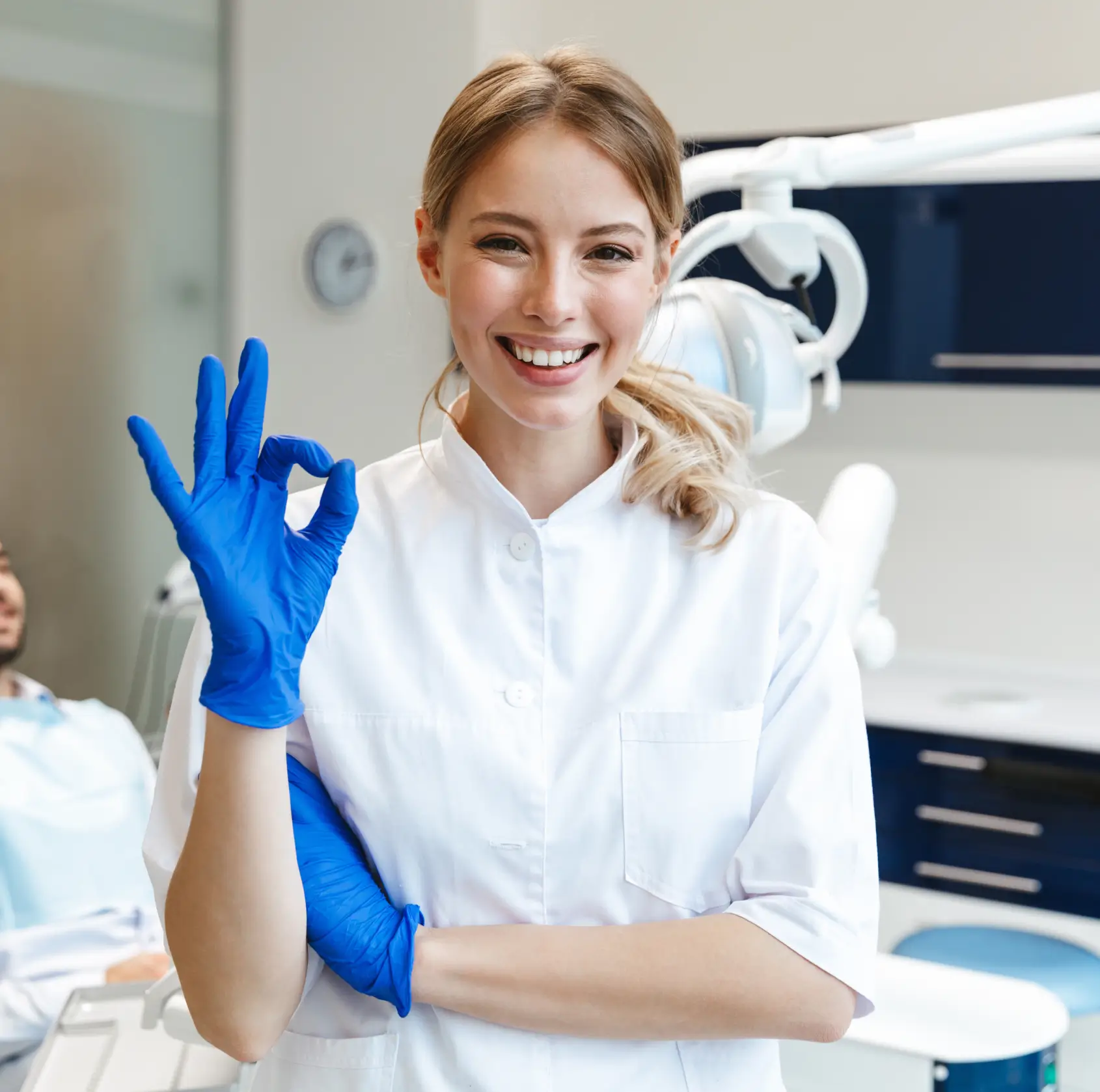 Smiling female dentist in white coat showing OK gesture with blue gloves in dental clinic. La Perle Dental Center in Edmonton, AB TST 3T9