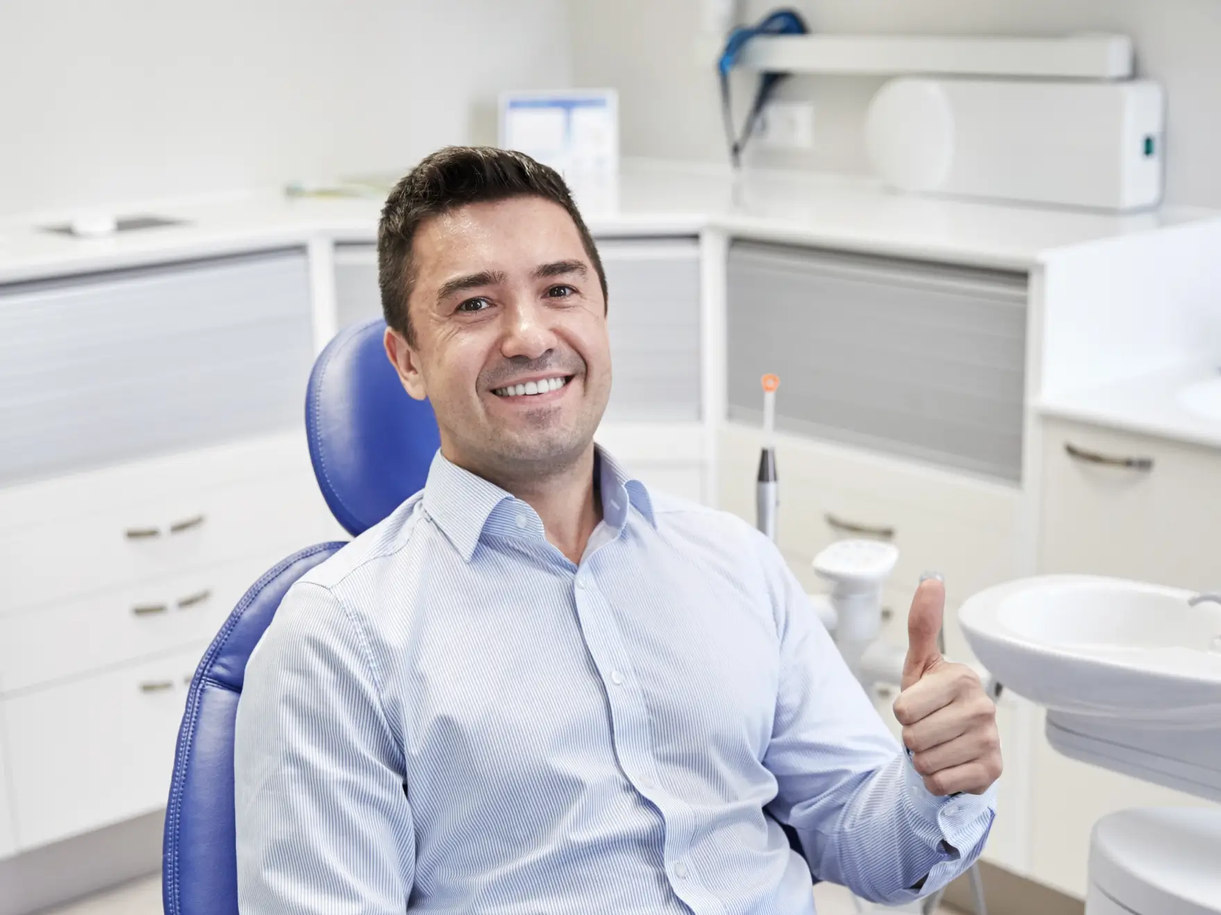 Smiling man in a blue shirt giving thumbs up while sitting in a dental chair at a clinic. La Perle Dental Center in Edmonton, AB TST 3T9