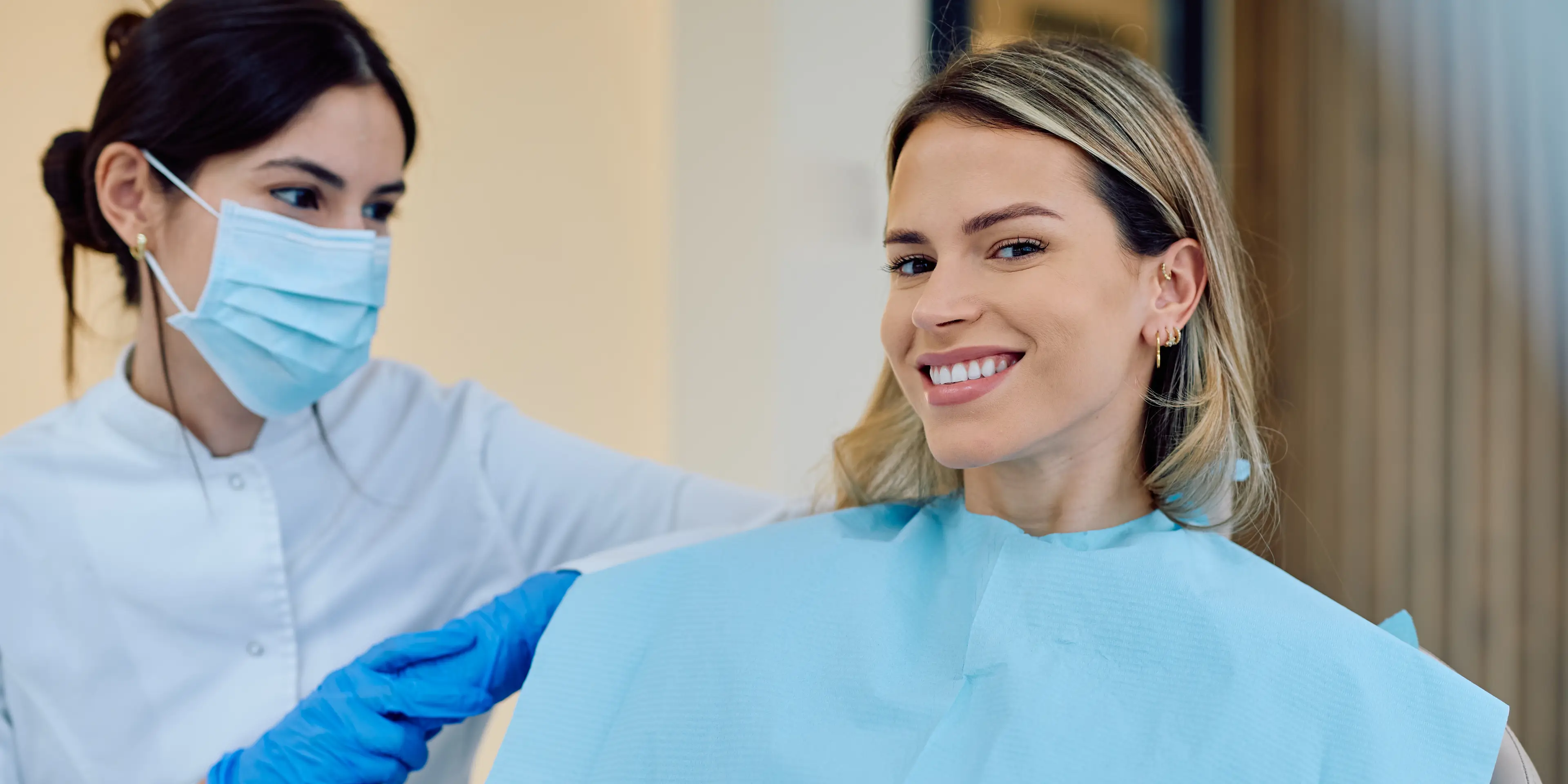 Smiling blonde woman in dental chair with blue bib, assisted by masked dental professional in gloves. La Perle Dental Center in Edmonton, AB TST 3T9