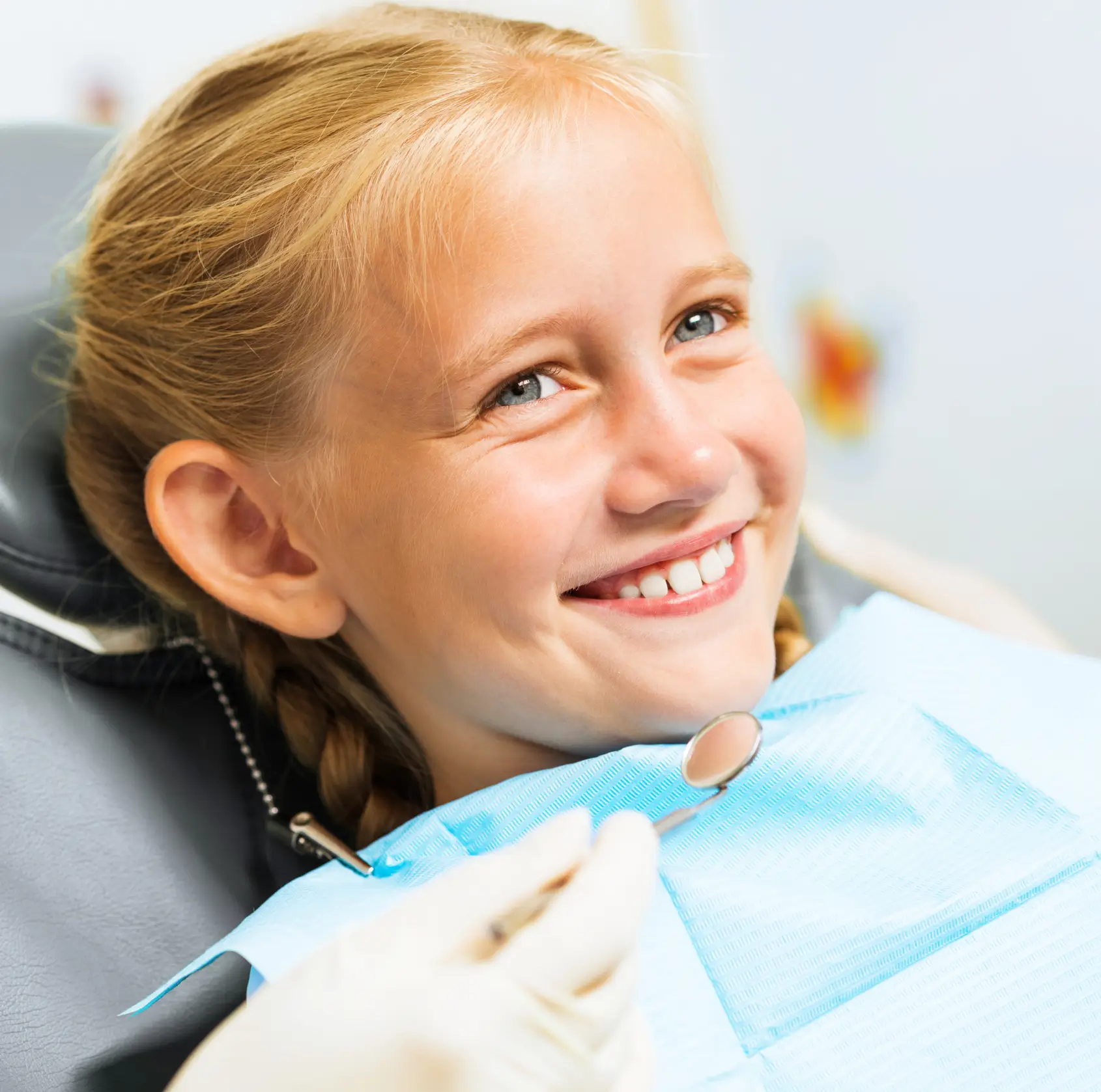 Smiling young girl sitting in a dental chair with a dental mirror held near her face. La Perle Dental Center in Edmonton, AB TST 3T9