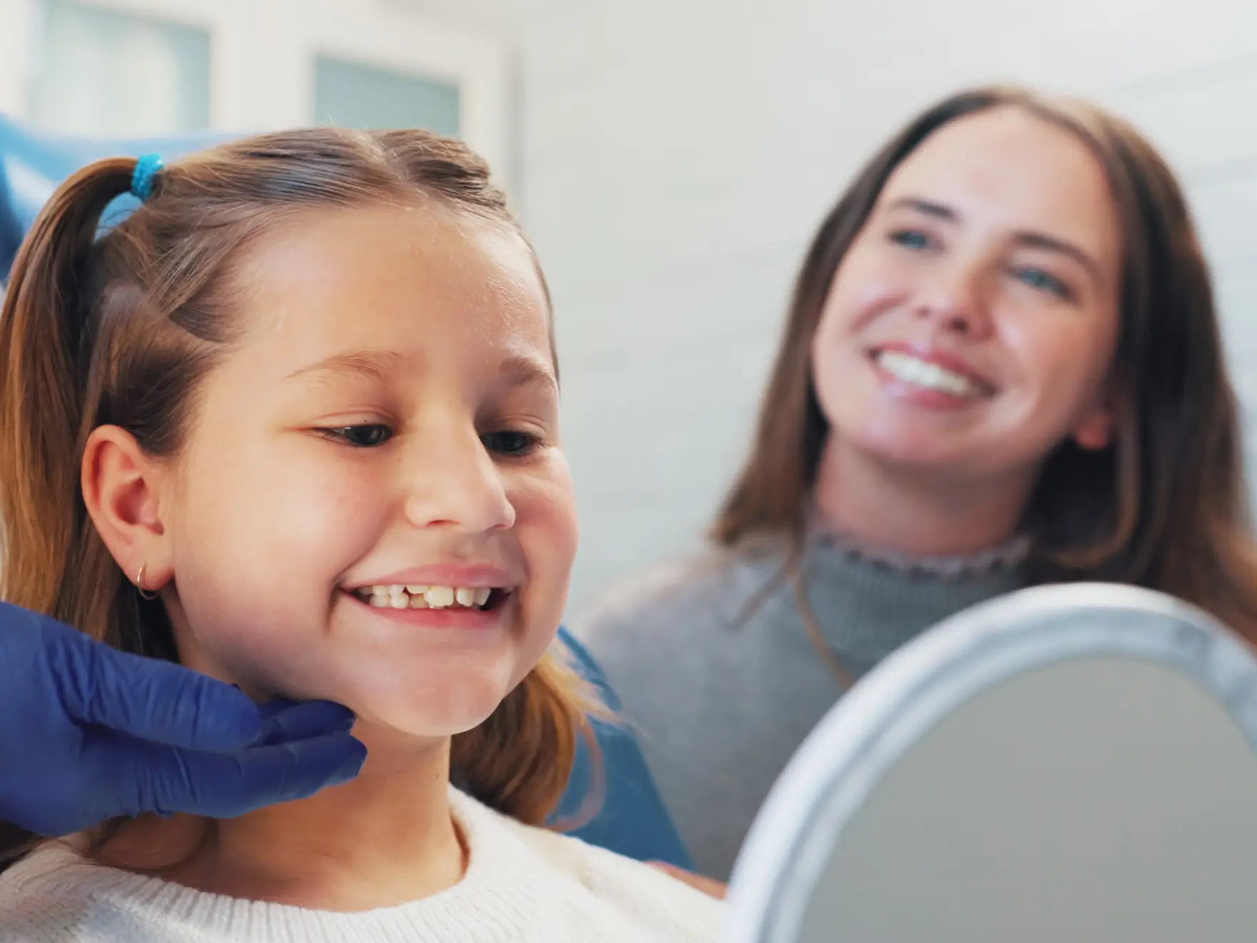 Smiling girl with pigtails checking her teeth in a mirror while a woman watches happily behind. La Perle Dental Center in Edmonton, AB TST 3T9