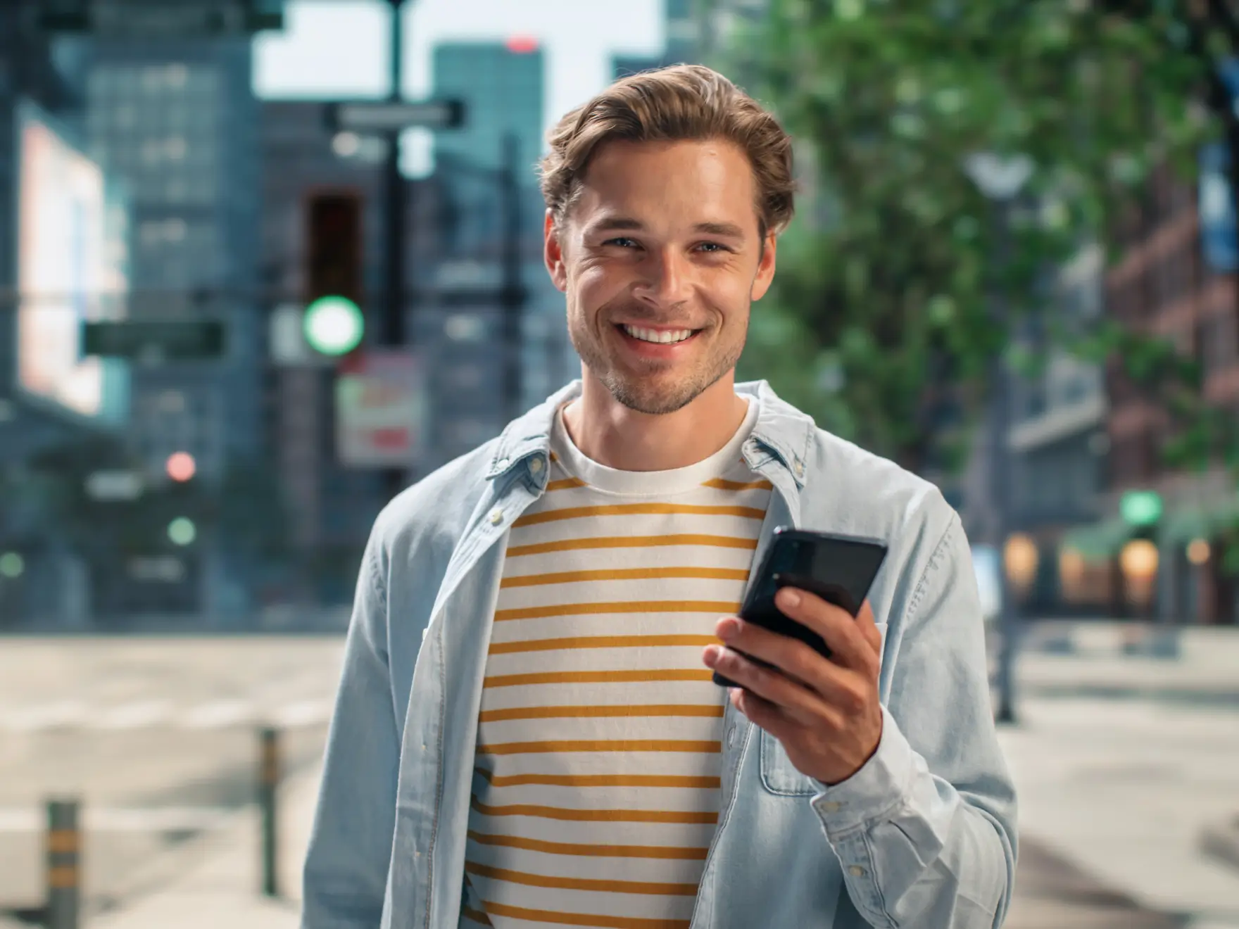 Smiling young man wearing striped shirt and denim jacket holding a smartphone outdoors in a city. La Perle Dental Center in Edmonton, AB TST 3T9